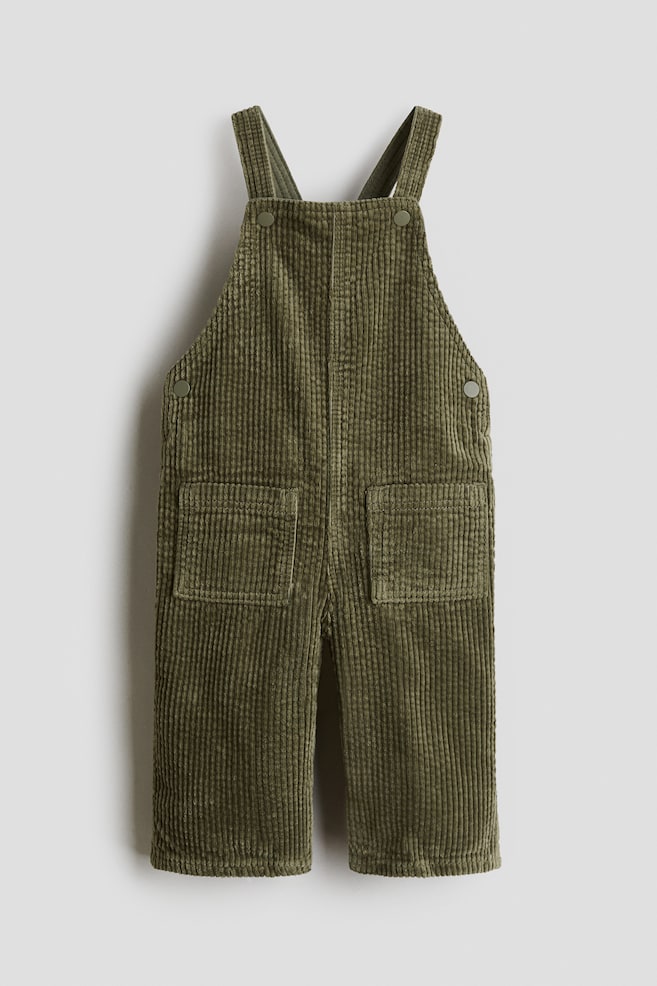 Une salopette en velours côtelé vert olive est présentée de face, avec des bretelles réglables boutonnées sur le plastron, des fermetures à boutons sur les côtés à la taille, et deux grandes poches plaquées à l'avant du pantalon. - 1