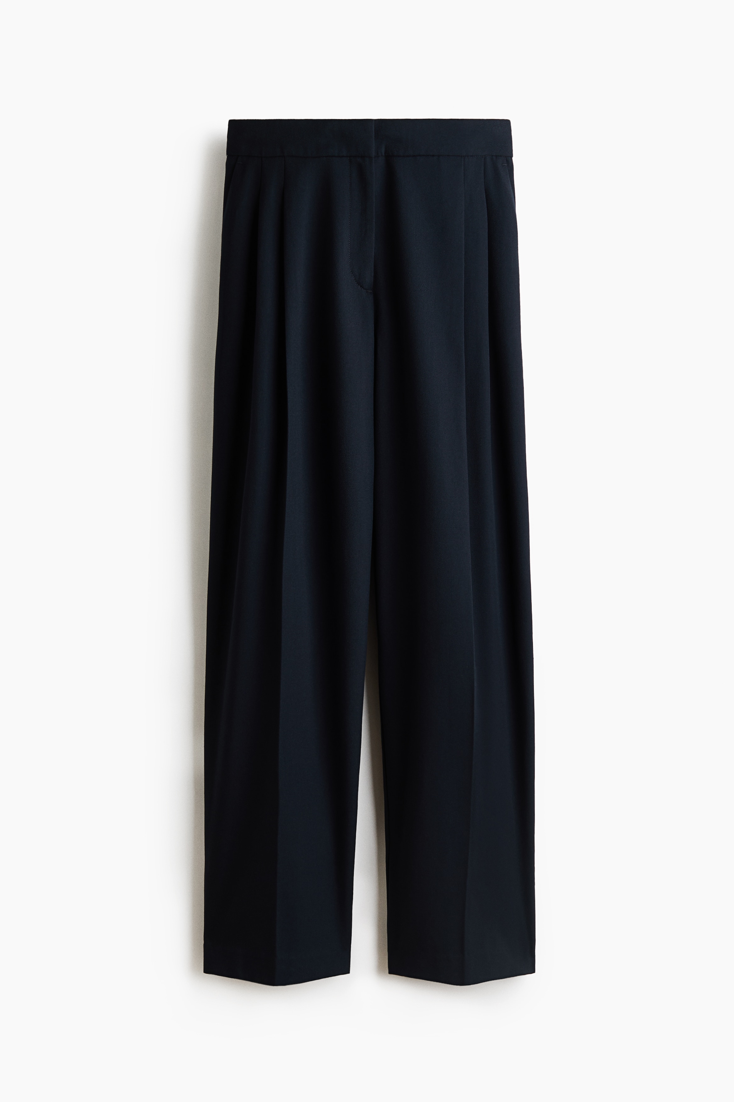 Tailored, loose-fit pants in dark navy are being worn by the model, who is standing with hands resting inside the side pockets. The high-waisted pants feature front pleats and creases down the barrel legs, styled with a tucked-in white short-sleeved top and black heeled sandals.