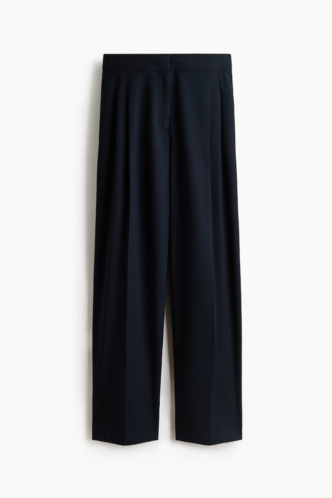 A pair of dark navy, loose-fit tailored pants is hanging and facing front, showcasing a high waist, pleats at the front, and sharp creases running down the wide, barrel-shaped legs. - 1