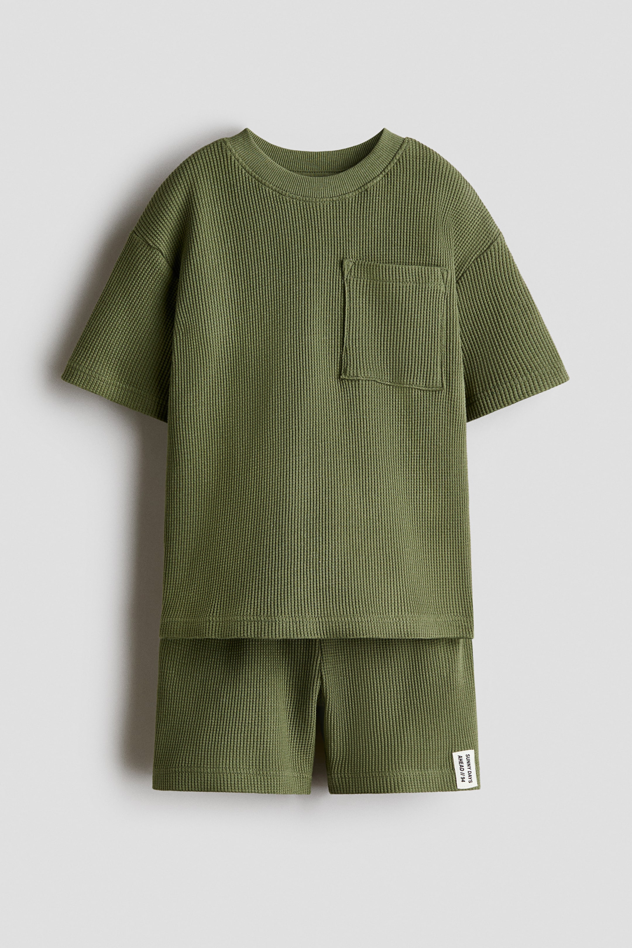 View larger image: An olive green waffled cotton jersey set is featuring a T-shirt and matching shorts, presented from the front. The T-shirt is displaying a round, rib-trimmed neckline, dropped shoulders, and a patch chest pocket, and the shorts are showing a small woven label on the right leg.