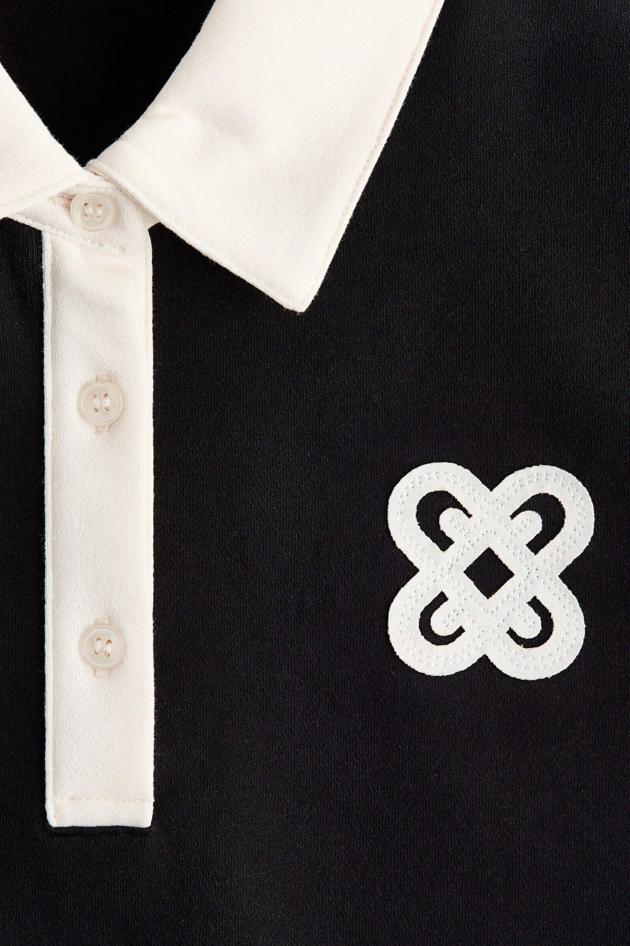 Xem ảnh lớn hơn: Một chiếc áo polo đen có cổ áo polo màu trắng ngà và hàng cúc ba nút phù hợp, với logo thêu màu trắng giống nút thắt nổi bật ở phía bên phải của ngực.