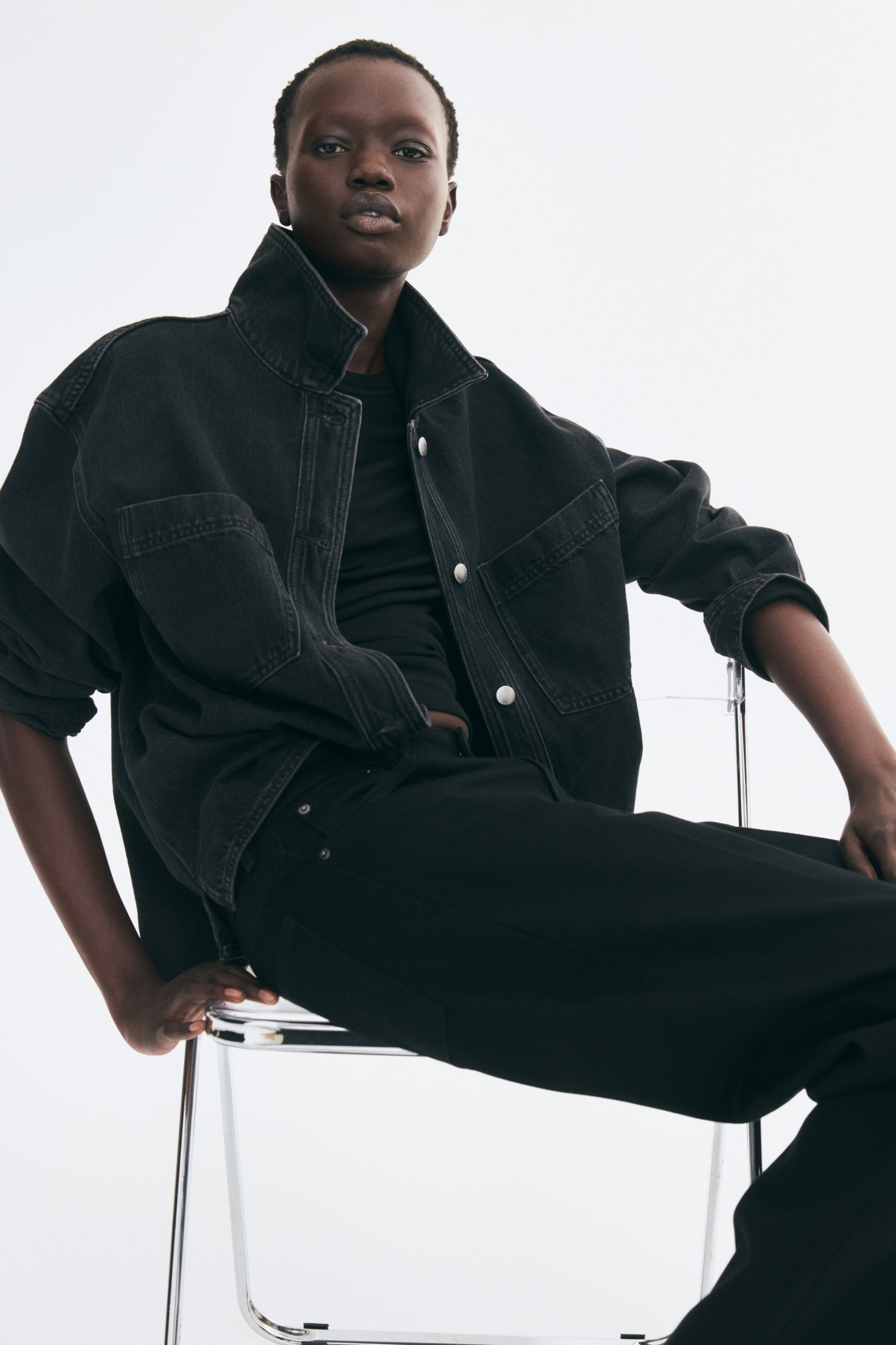 Grotere afbeelding bekijken: Een oversized, zwart gewassen denim overshirt wordt open gedragen door het model, waardoor de losse pasvorm, kraag, verlaagde schouders en twee open borstzakken mooi zichtbaar zijn. Het overshirt wordt gedragen over een zwart T-shirt en gecombineerd met een zwarte broek.
