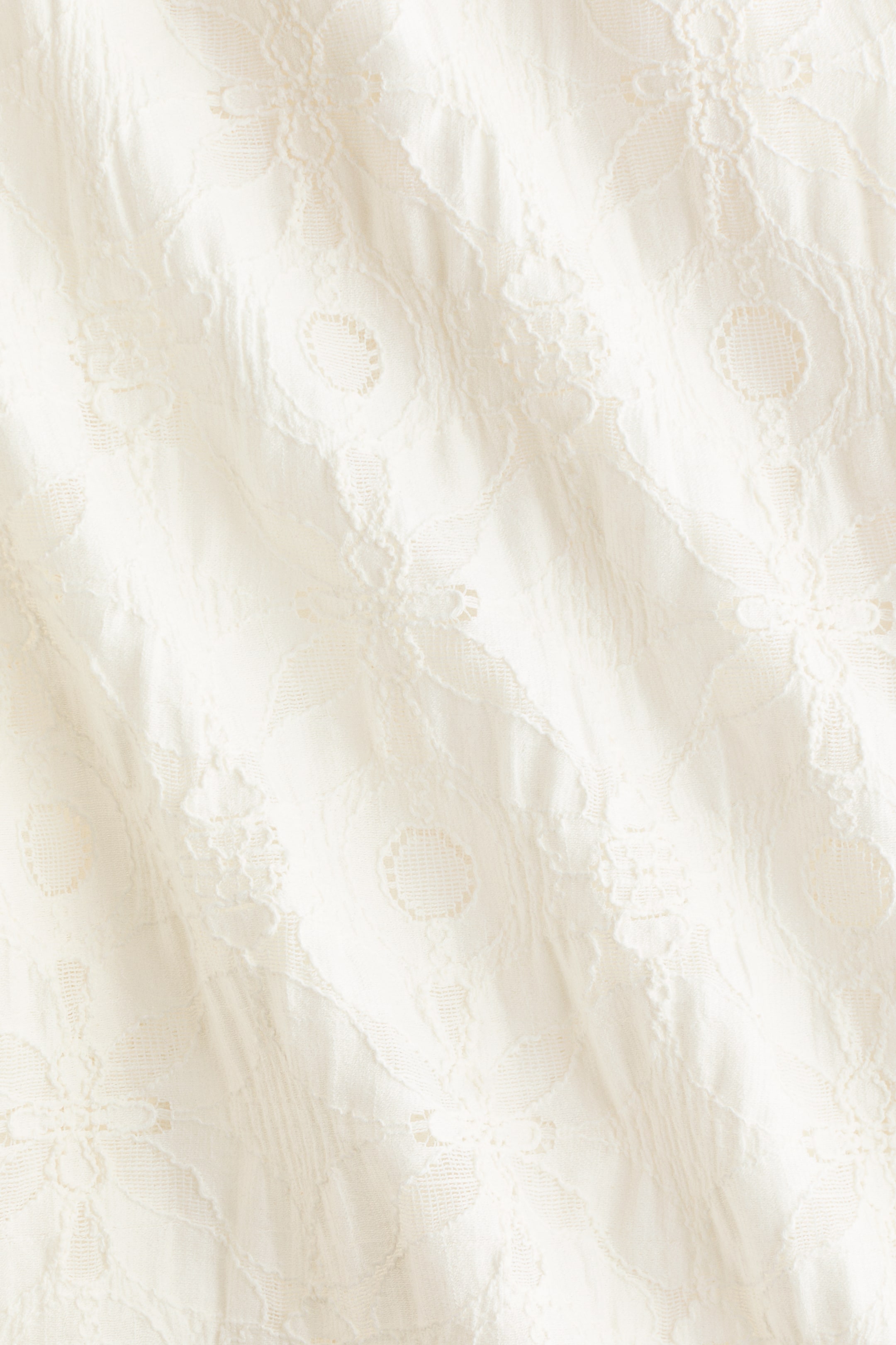 Visualizza immagine più grande: Pantaloni a vestibilità ampia in jersey effetto pizzo sono mostrati in primo piano, concentrandosi sul tessuto color crema. Il tessuto presenta un motivo intricato e in rilievo di motivi floreali e circolari, mostrando morbide pieghe.