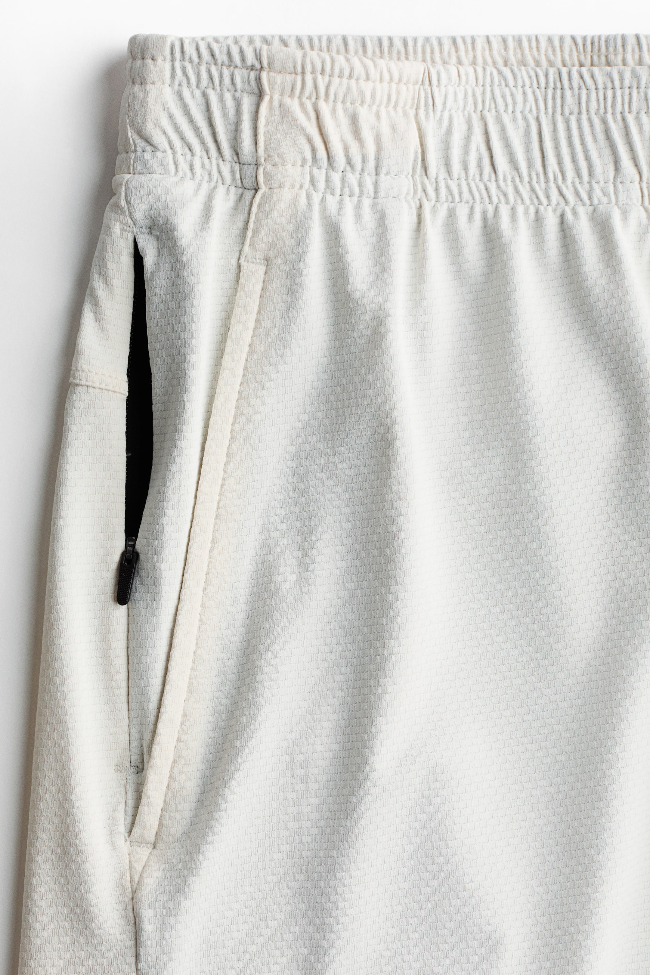 Ampliar la imagen: Un par de shorts deportivos de color claro presentan una pretina elástica, una textura de cuadrícula sutil y un bolsillo con cremallera negra en ángulo en el lado izquierdo con un tirador de cremallera negra visible y un cordón.