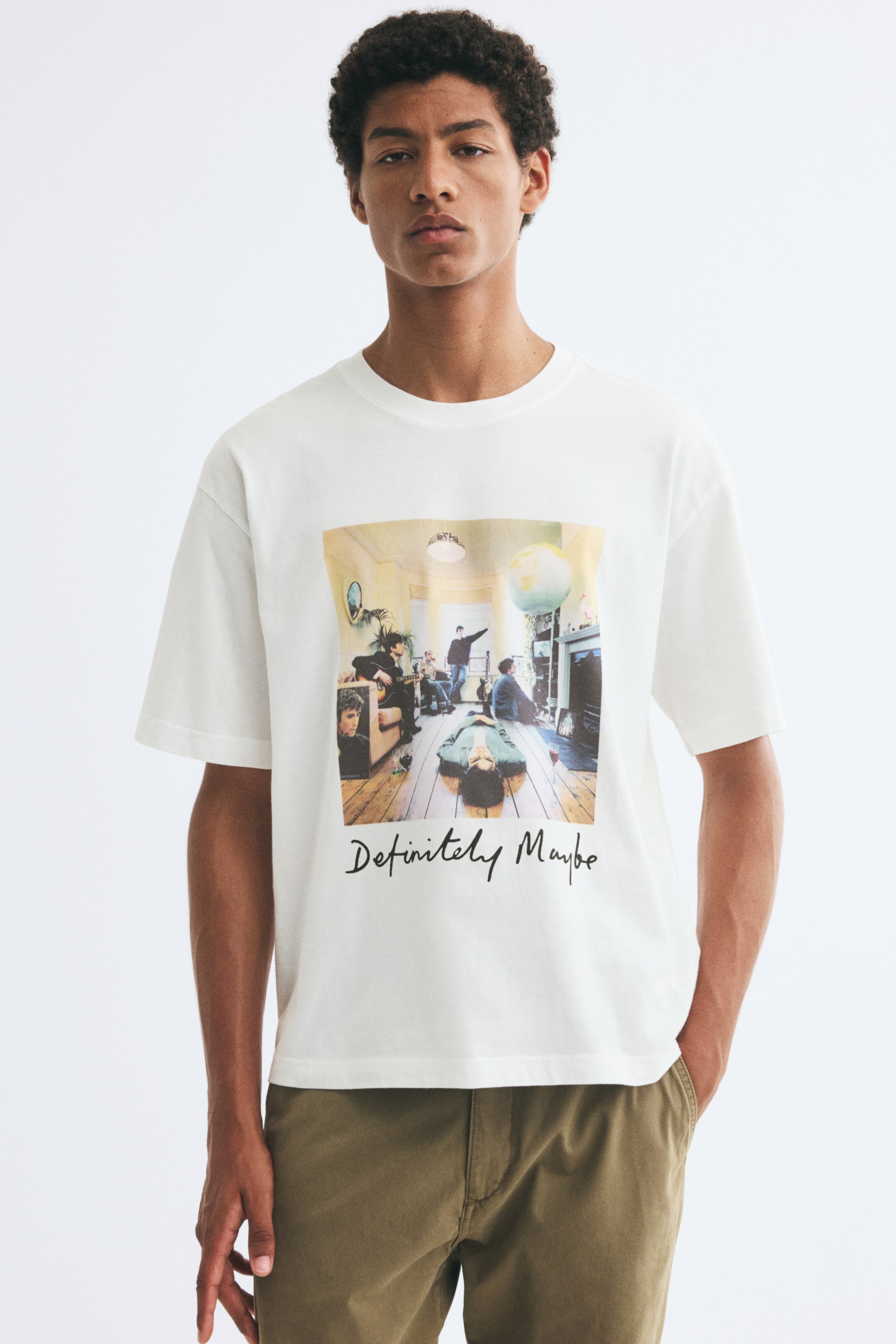 Größeres Bild ansehen: Ein locker geschnittenes T-Shirt in Off-White mit geripptem Rundhalsausschnitt, überschnittenen Schultern und einem Front-Print des Albumcovers von „Definitely Maybe“ wird vom Model getragen. Das T-Shirt ist mit olivgrünen Hosen kombiniert.