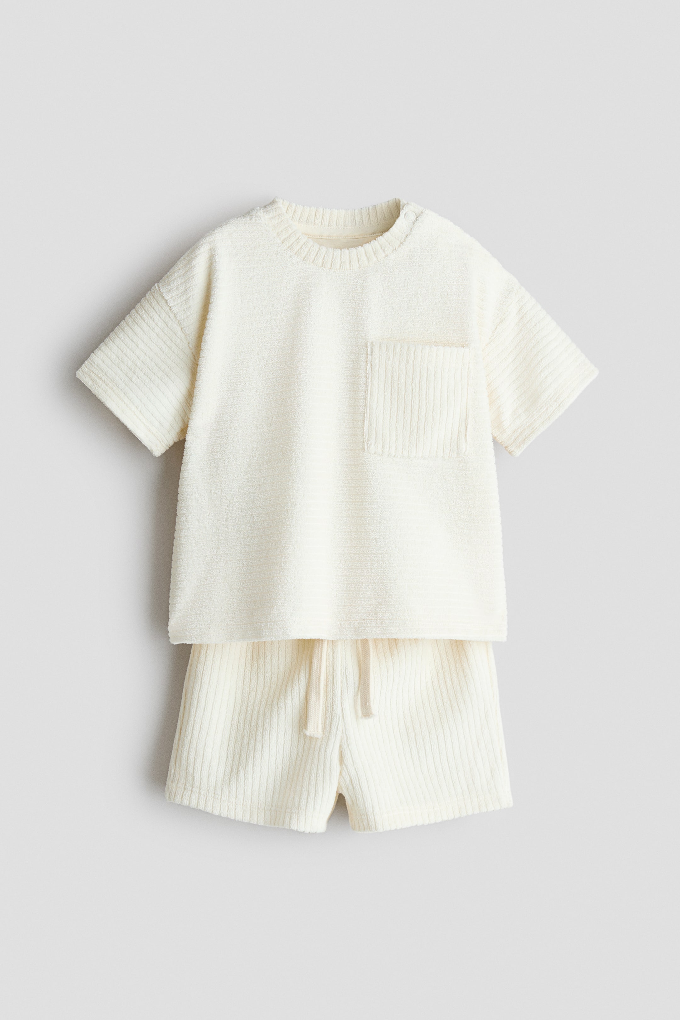 Prikaži veću sliku: Dvodelni rebrasti set od frotira predstavlja krem majicu kratkih rukava sa okruglim izrezom i odgovarajuće šorts, prikazane s prednje strane. Majica ima rebrasti okrugli izrez, kratke rukave sa horizontalnim rebrastim detaljima i vertikalni rebrasti džep na levom delu grudi; glavni deo majice i šorts prikazuju vertikalne rebraste detalje, dok šorts takođe ima pojas sa uzicom.