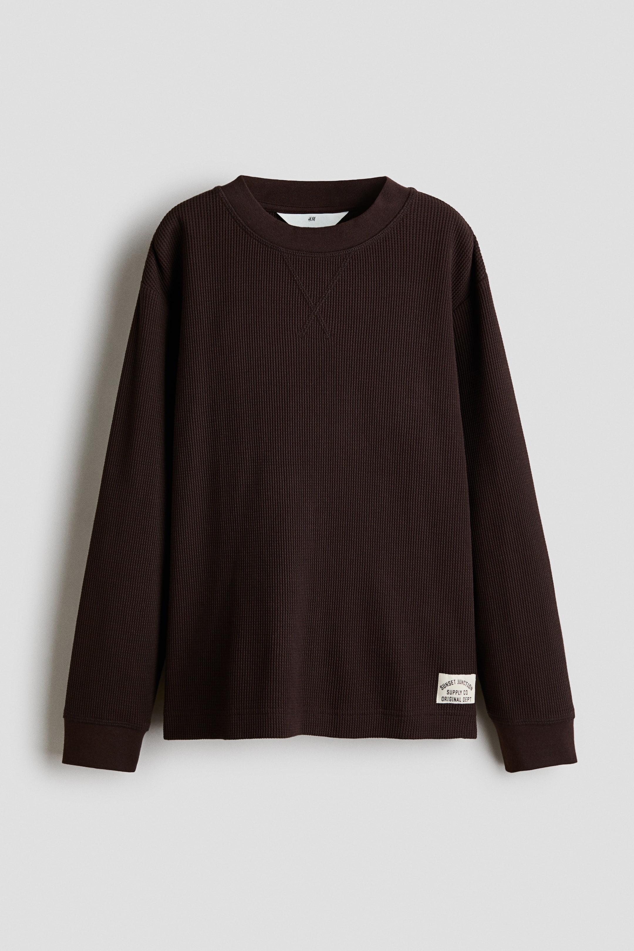 View larger image: A dark brown waffle-knit long-sleeved top is shown from the front, featuring a ribbed crew neck with V-shaped stitching below the collar, ribbed cuffs, and a ribbed hem. A small rectangular fabric label with text is sewn onto the lower left front.