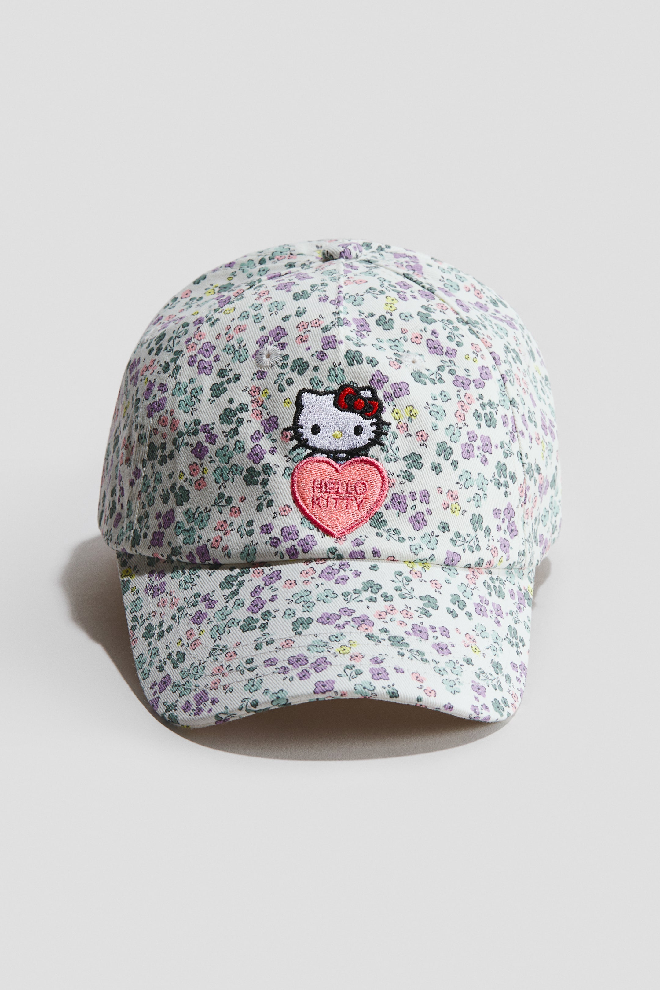 View larger image: A cap is facing front, showcasing a dense, petite floral print in shades of purple, green, pink, and yellow on a light background. An embroidered patch is centrally positioned on the front, depicting Hello Kitty with a red bow above a pink heart that reads 'HELLO KITTY'.