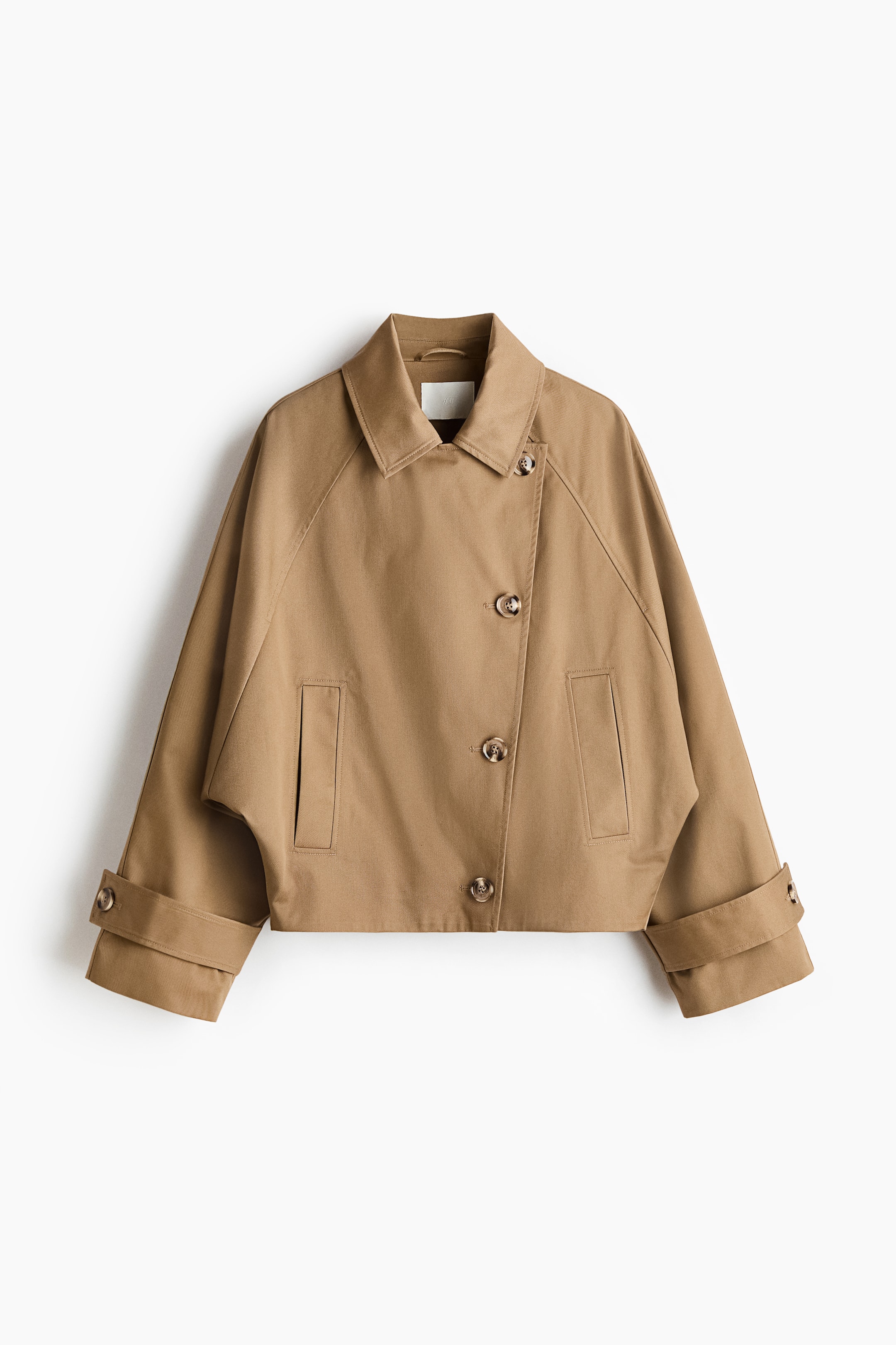 View larger image: A tan, short, oversized trench coat is showcasing its front. It features a classic collar, an off-set button placket with multiple buttons, two horizontal welt pockets, and raglan batwing sleeves with tab and button cuffs.