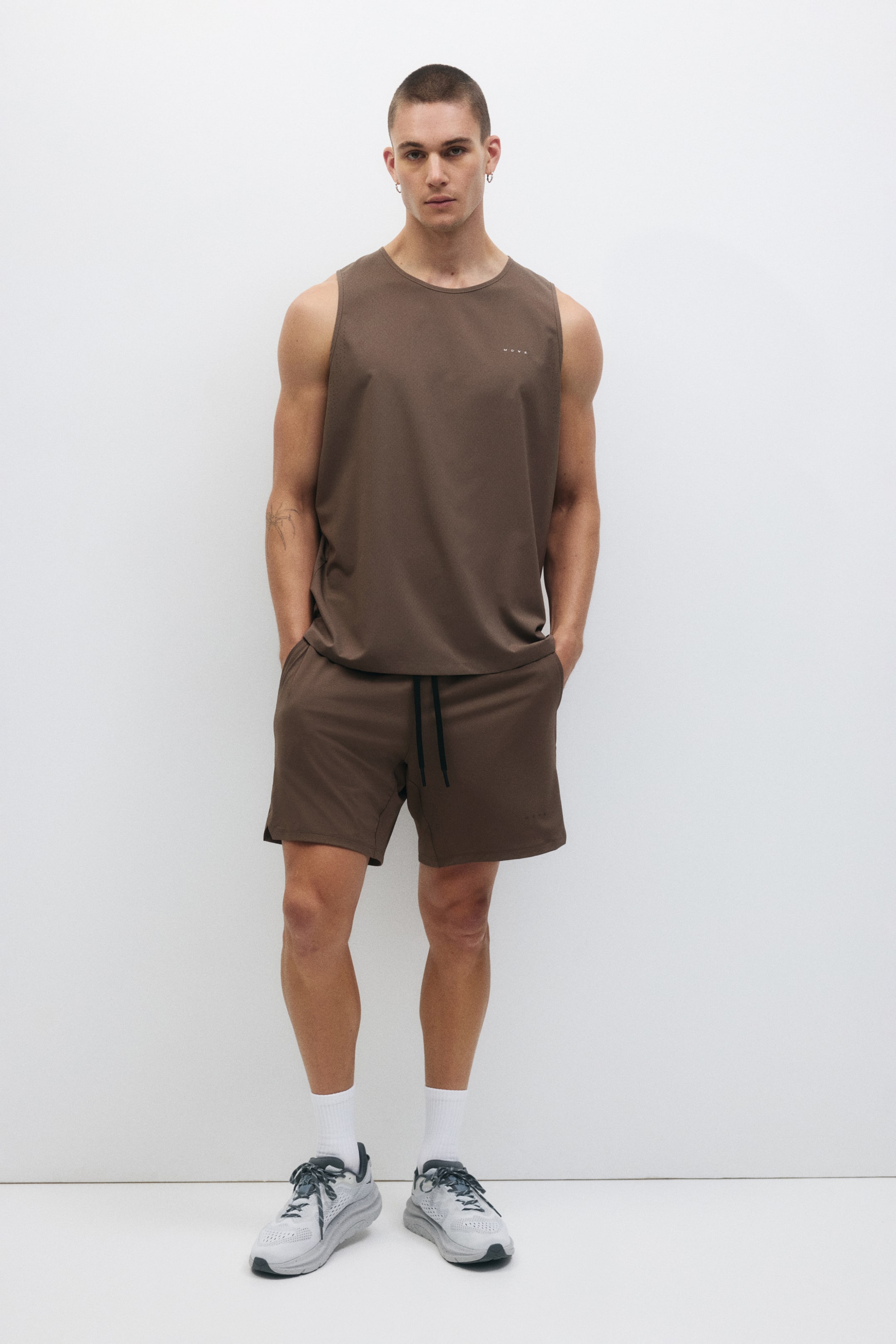 Visa större bild: Ett brunt ärmlöst linne och matchande shorts presenteras som ett koordinerat set, vända framåt. Linnet har en rund hals och en liten vit logotyp på vänster bröst, medan shortsen har en elastisk midja med ett svart dragsnöre och sidofickor.