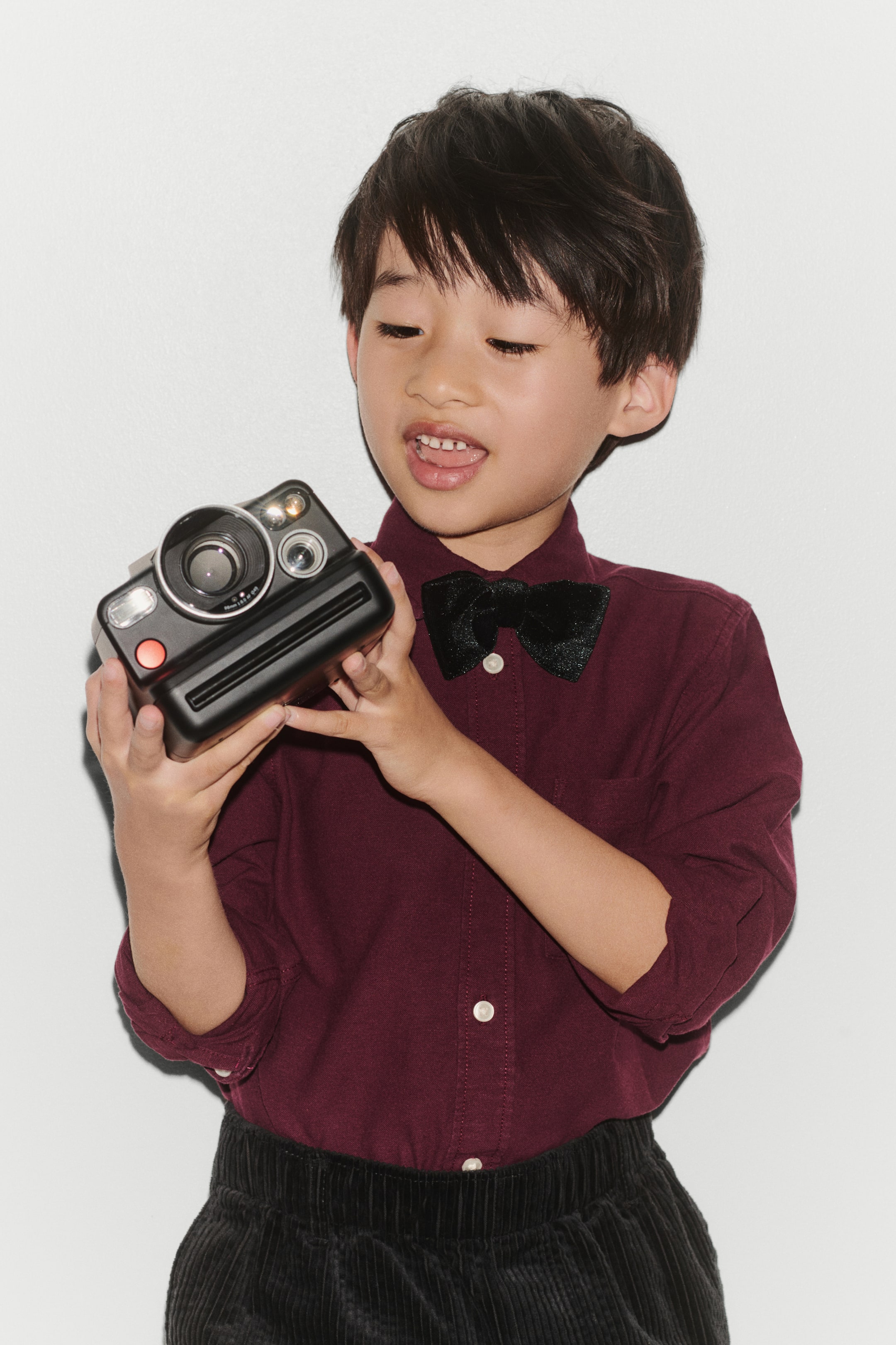 View larger image: A burgundy Oxford cotton shirt is worn in a front view, featuring a turn-down collar styled with a black bow tie and a button-down front with white buttons. The shirt has long sleeves pushed up and is tucked into black corduroy trousers.