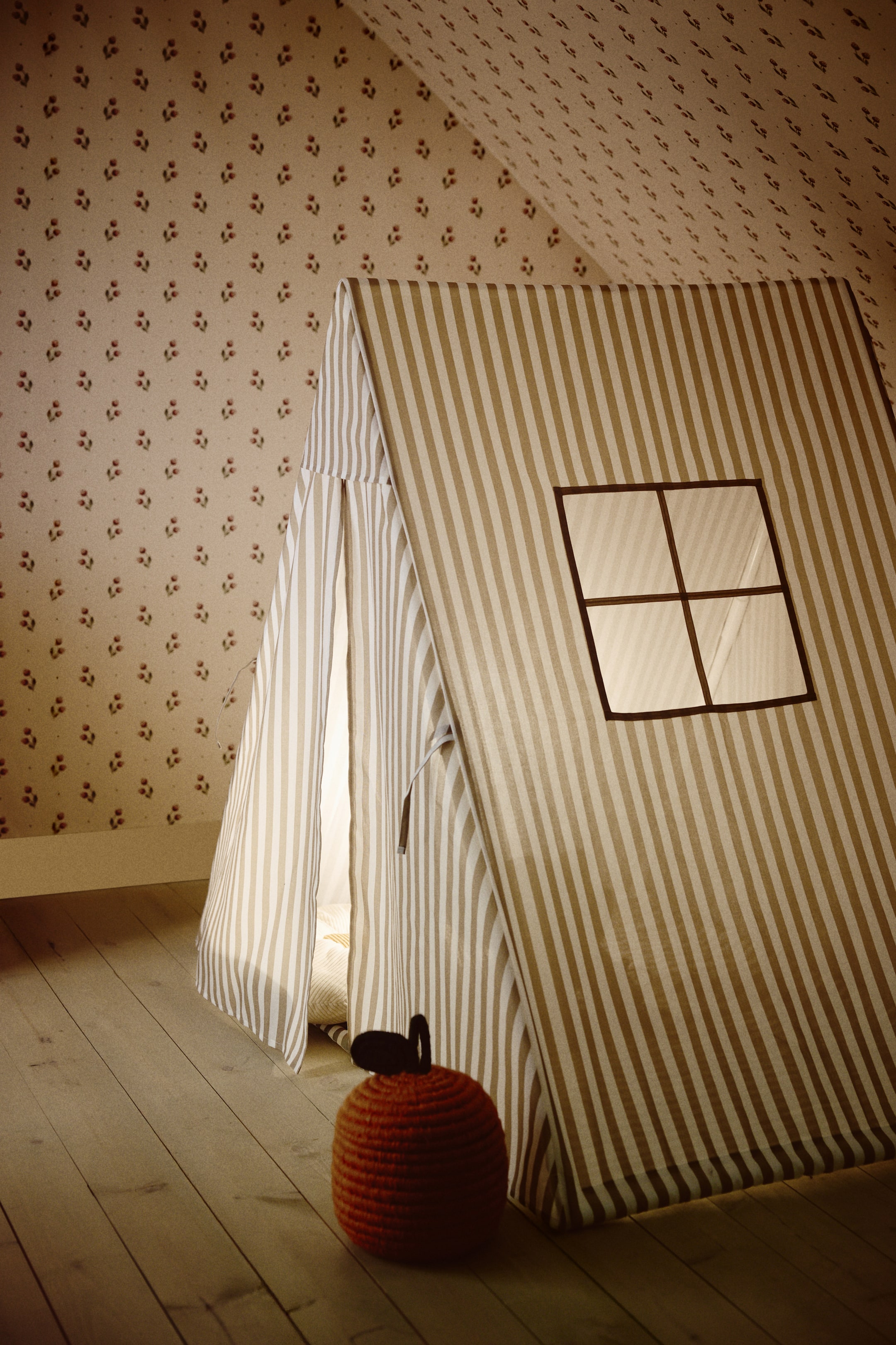 Nagyobb kép megtekintése: Egy játék sátor enyhén megdőlve áll, függőlegesen váltakozó világos és sötét bézs csíkokkal, bal oldalon részben nyitott textil bejárattal, valamint a jobb elülső panelen egy sötét keretes, keresztpántos négyzet alakú ablakkal. Egy piros kötött alma pihen a sátor előtt a padlón.