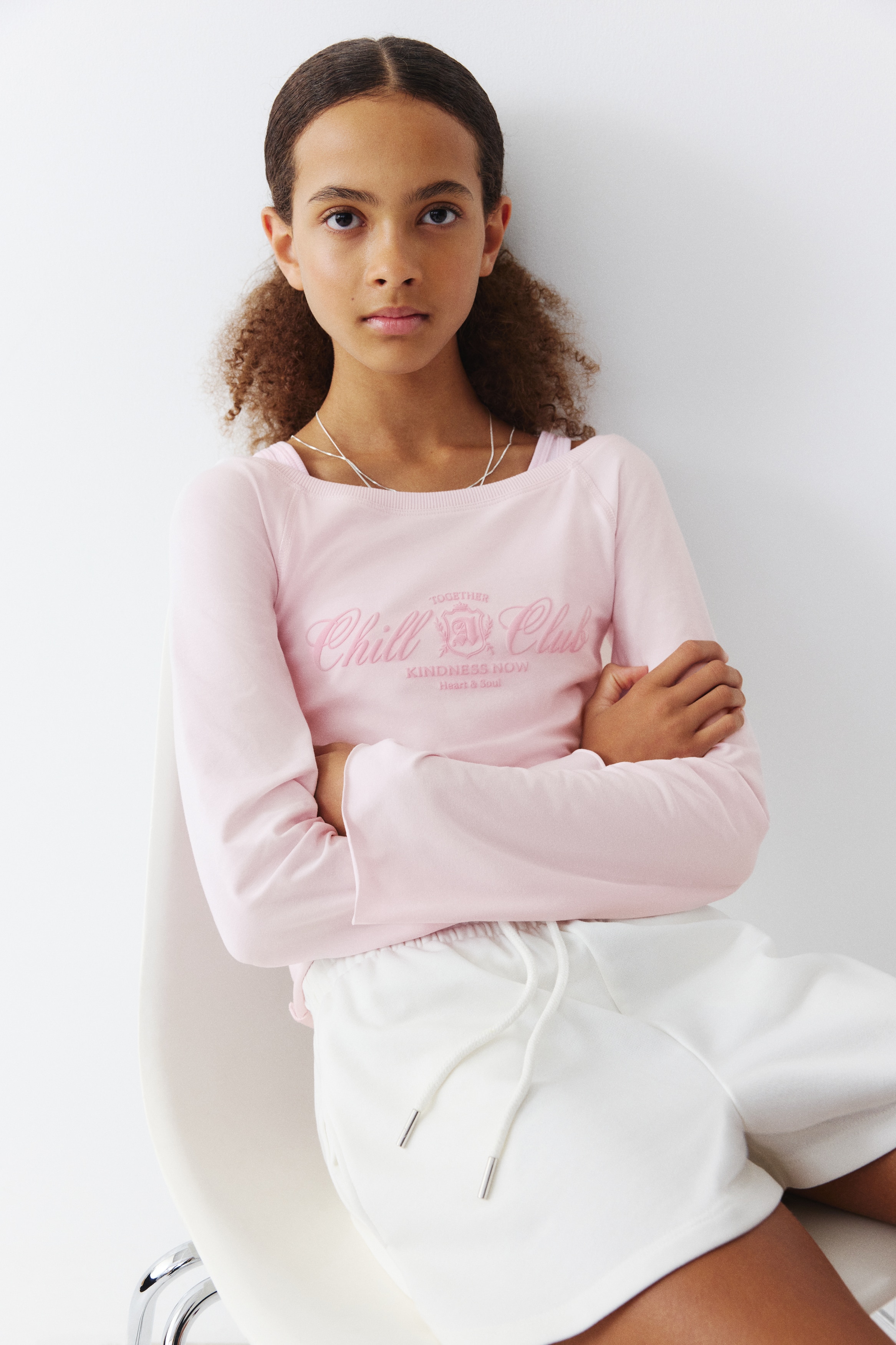 This front view shows a light pink sweatshirt top featuring a wide boat neckline with rib trim and wide raglan sleeves. The chest features a pink text print reading "Chill Club," while the cuffs and hem feature raw, rolled-up edges.