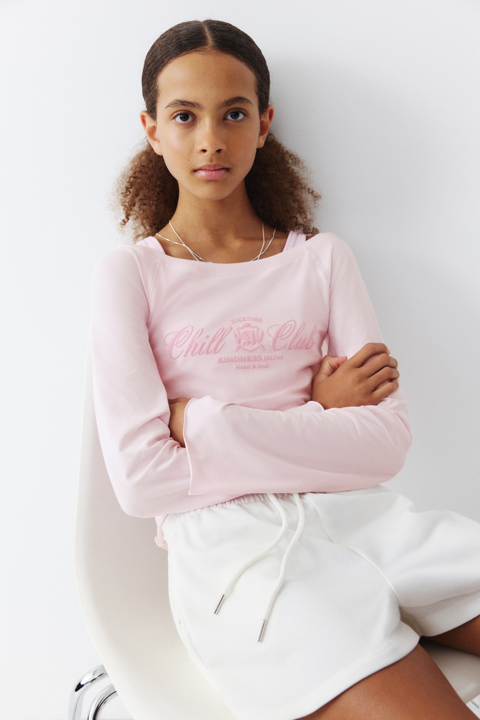 This front view displays a light pink sweatshirt top featuring a wide, rib-trimmed boat neckline and wide raglan sleeves. The chest shows a pink text print that reads "Chill Club," while the cuffs and hem feature raw, roll-up edges. - 1