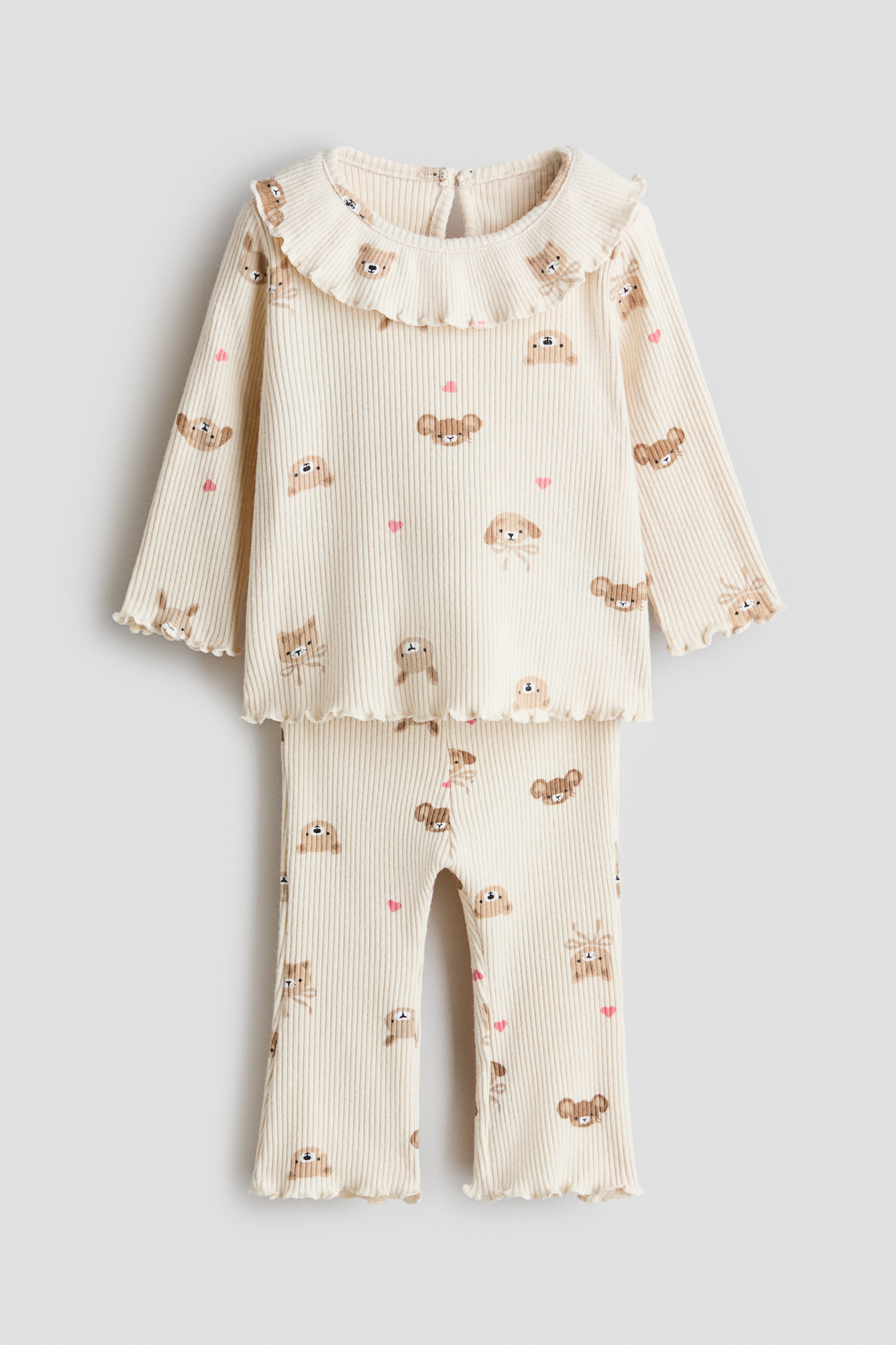 View larger image: A FRONT view shows a cream-colored, ribbed two-piece pajama set for children, featuring an all-over print of various brown animal faces and small pink hearts. The long-sleeved top features a ruffled collar, a keyhole opening at the back, and lettuce-edged cuffs and hem, while the matching long pants display lettuce-edged leg openings.