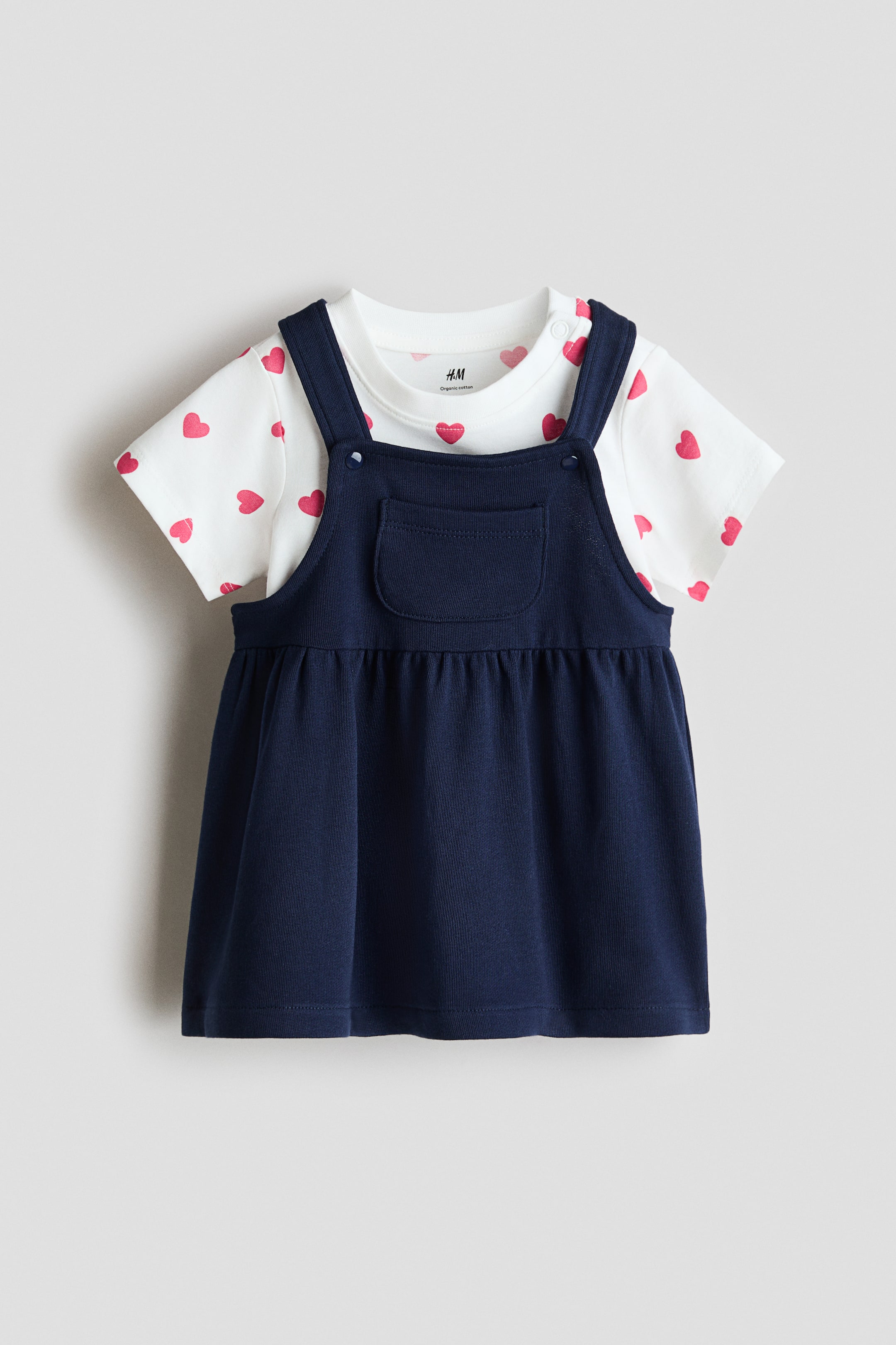 View larger image: A baby outfit displays a white, short-sleeved top with a red heart print, layered under a navy-blue pinafore dress. The pinafore, seen from the FRONT, features shoulder straps with buttons, a small pocket on the bib, and a gathered skirt.
