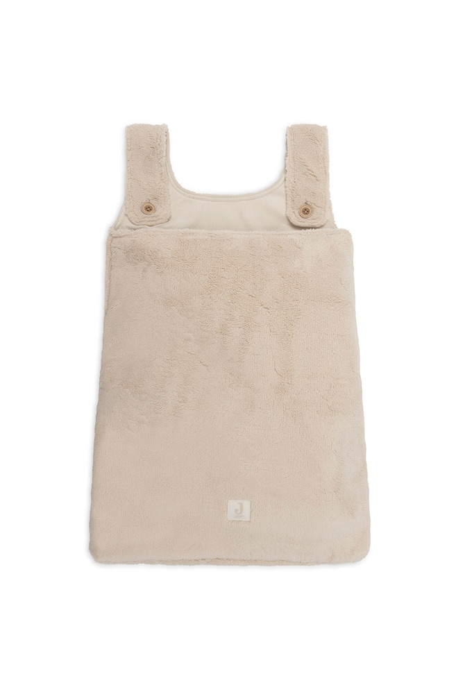 Un sac de couchage pour bébé en peluche beige est posé à plat et orienté vers l'avant, mettant en valeur sa texture en polaire douce et duveteuse. Le modèle sans manches présente de larges bretelles maintenues à l'avant par de simples boutons marron, ainsi qu'un petit patch logo carré cousu près du centre inférieur. - 1