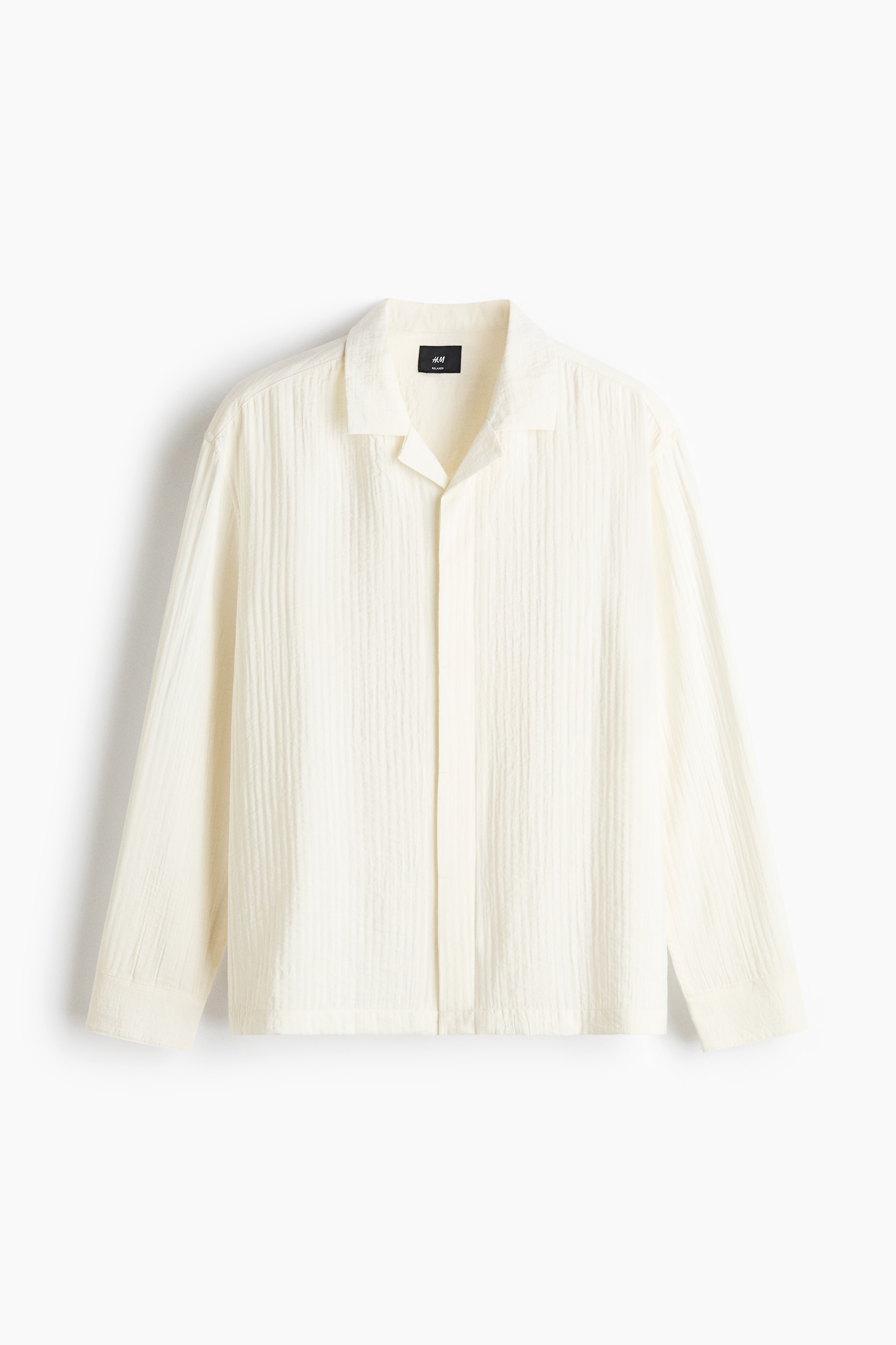 An off-white shirt in an airy, textured weave is worn by the model, featuring a relaxed fit, a resort collar, and long sleeves with button cuffs. The shirt is styled untucked with loose-fitting beige trousers and dark sandals.