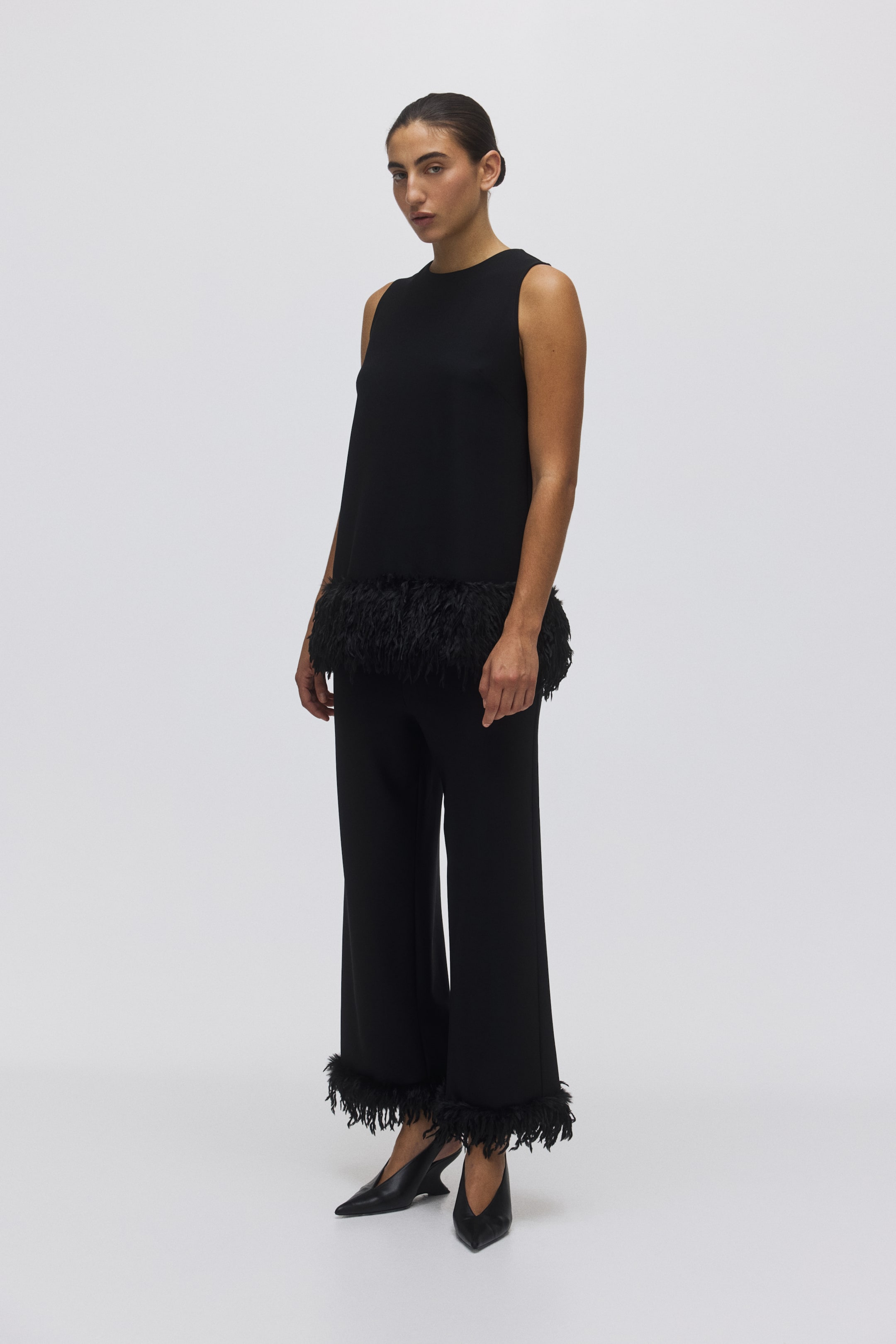 View larger image: A black sleeveless top and matching trousers are shown from a slightly angled front view. The top is featuring a round neck and a feather trim along its bottom hem. The trousers are designed with a wide leg and also feature a feather trim at the hems.