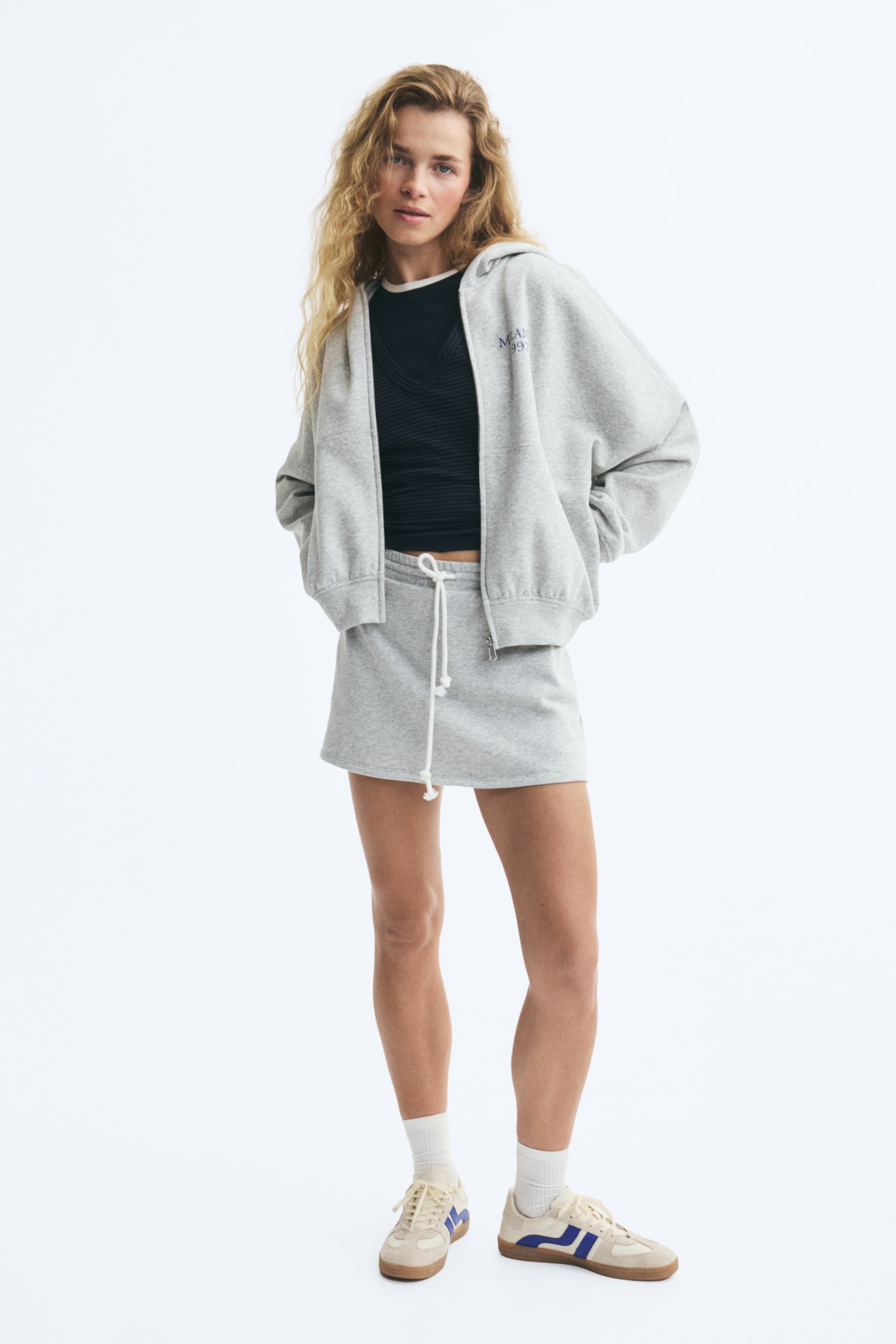 View larger image: A relaxed-fit, light grey fleece hoodie is worn unzipped by the model, featuring dropped shoulders, ribbed cuffs, and a small embroidered graphic on the chest. The hoodie is layered over a black top and paired with a matching light grey fleece skirt.