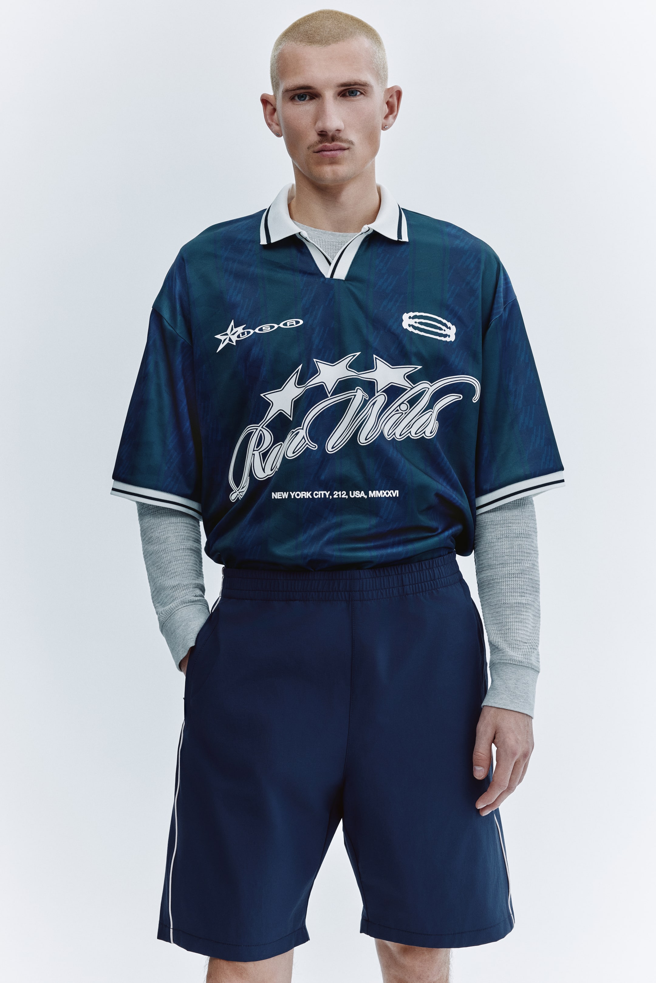 View larger image: A short-sleeve soccer jersey in dark teal and navy displays a striped pattern and white graphics, including a star-outline logo, "Raw Wild" script, and text, and features a white pointed collar and striped sleeve cuffs. It is worn layered over a light-gray long-sleeve thermal shirt and paired with dark navy athletic shorts with an elastic waistband and white side stripes.