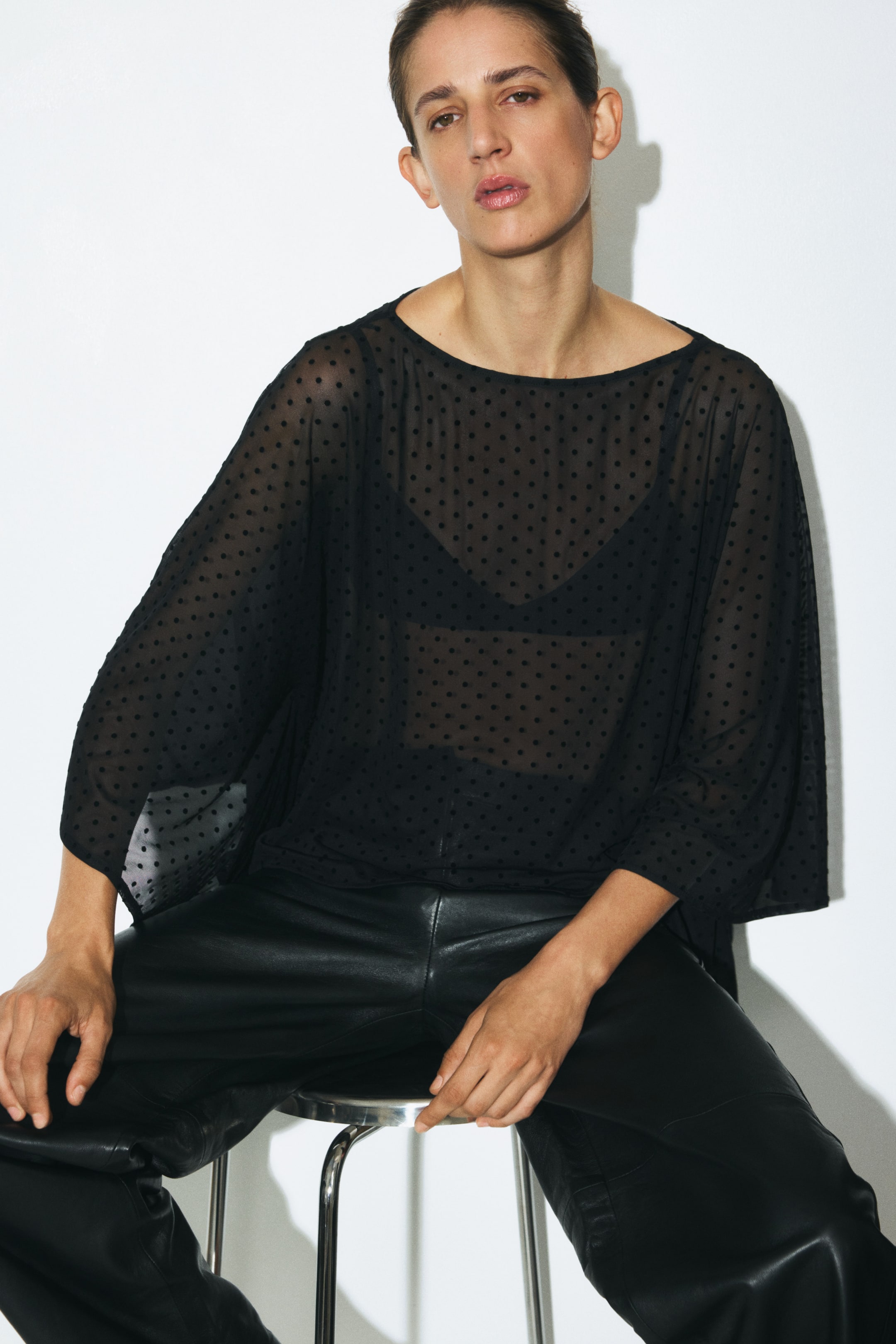 Ampliar la imagen: Una blusa tipo capa de corte oversized en tela negra transparente con lunares, con un delicado escote barco y bajo tipo pañuelo, la lleva la modelo sobre un bralette negro. La blusa se combina con pantalones negros de piel.