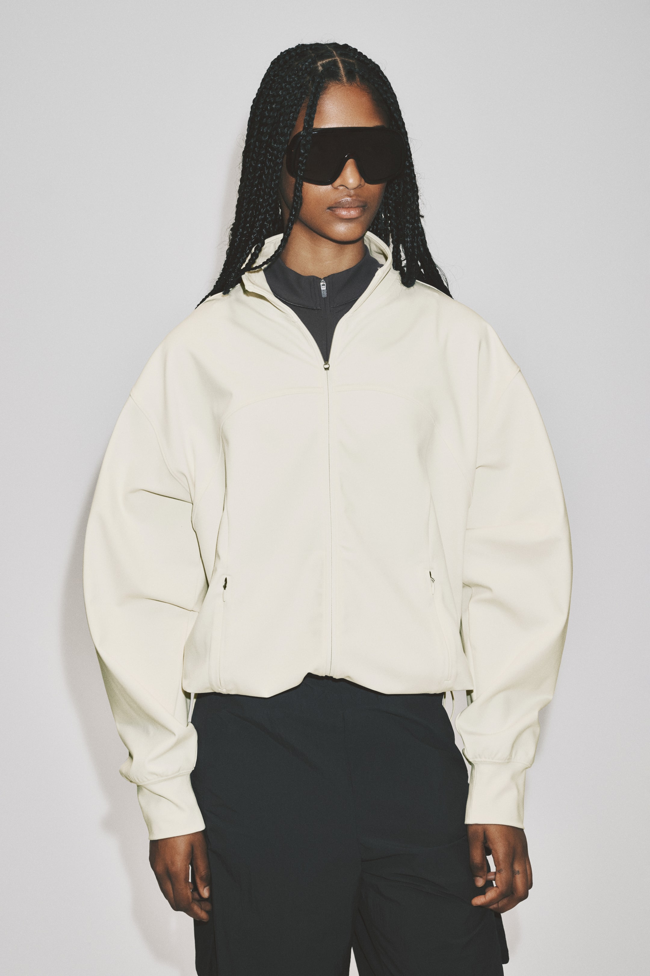 Ver imagen más grande: Chaqueta deportiva con SoftMove? La chaqueta de color crema, con cierre completo en la parte delantera, un cuello alto, dos bolsillos laterales con cremallera y puños y dobladillo elásticos, está siendo llevada por la modelo. La modelo la combina con un top negro de cuello alto y pantalones negros.