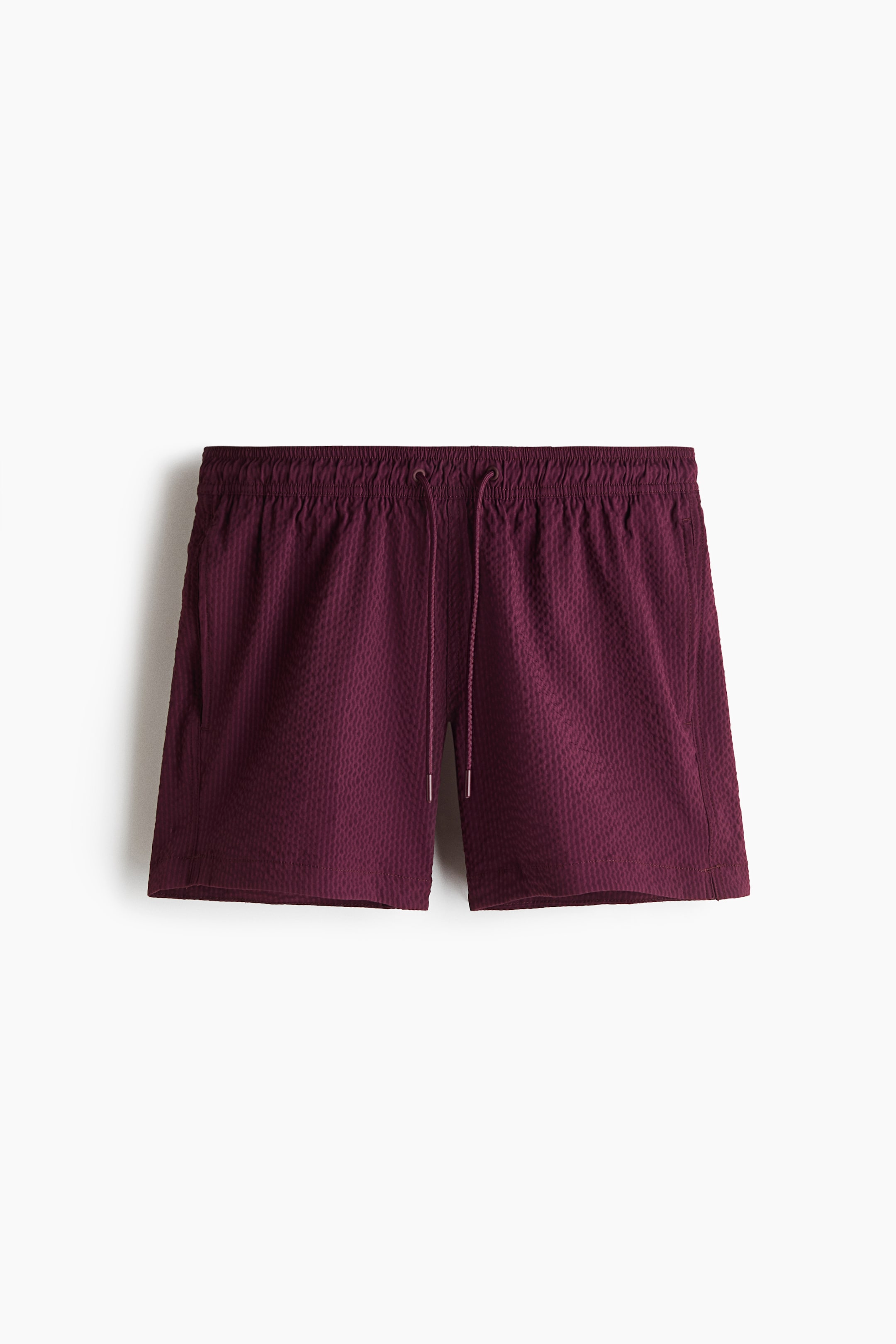 View larger image: Swim shorts in maroon seersucker fabric are shown from the front, featuring a covered elastic waistband with a white-tipped drawstring. Side pockets are visible, and small slits appear at each side of the hem.