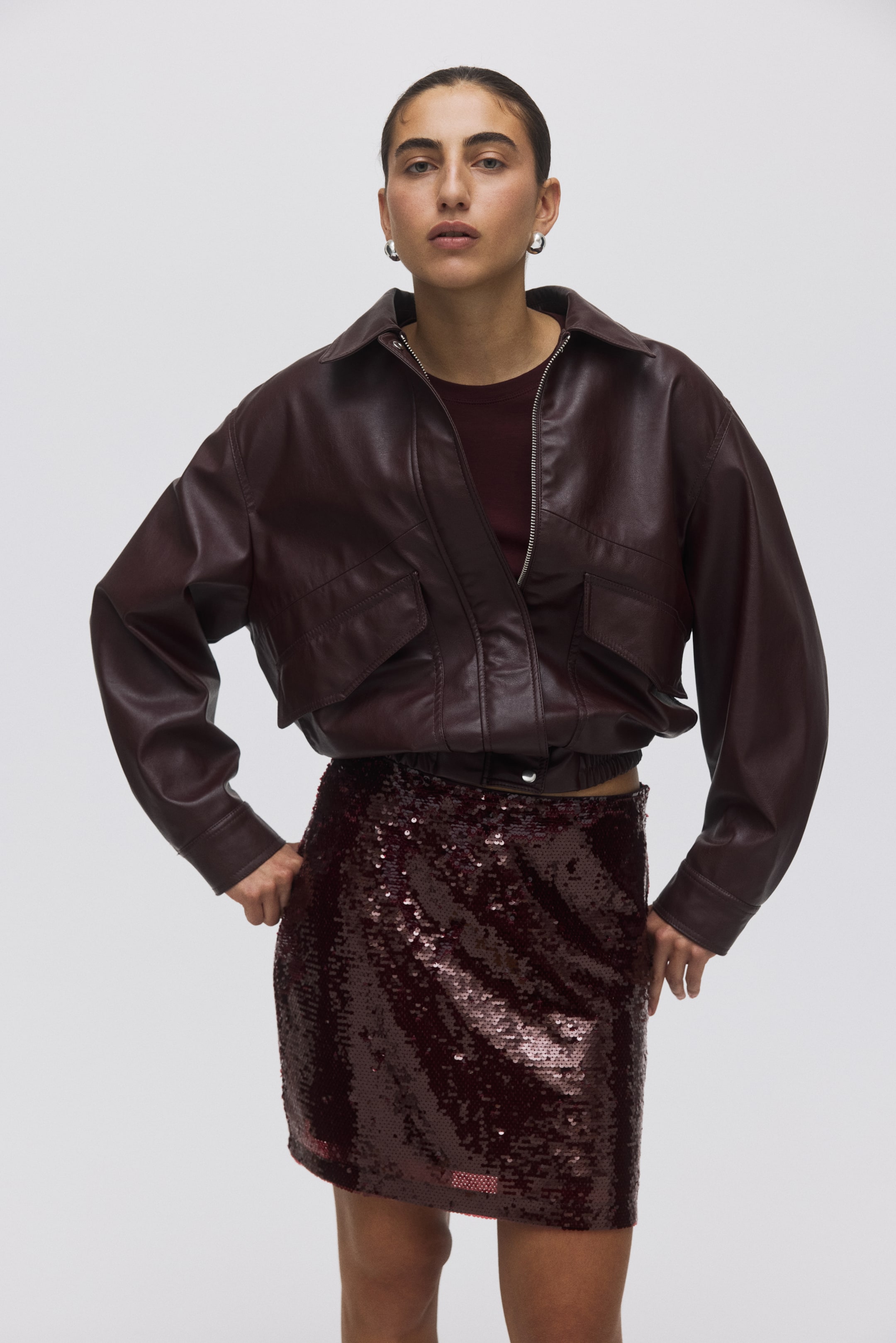 View larger image: A dark red faux leather bomber jacket is being presented from a front-facing angle, slightly to the right, showing a wide collar, a full-zip closure, two large flap pockets on the chest, and an elasticated hem and cuffs. It is being worn over a dark red crew-neck top.