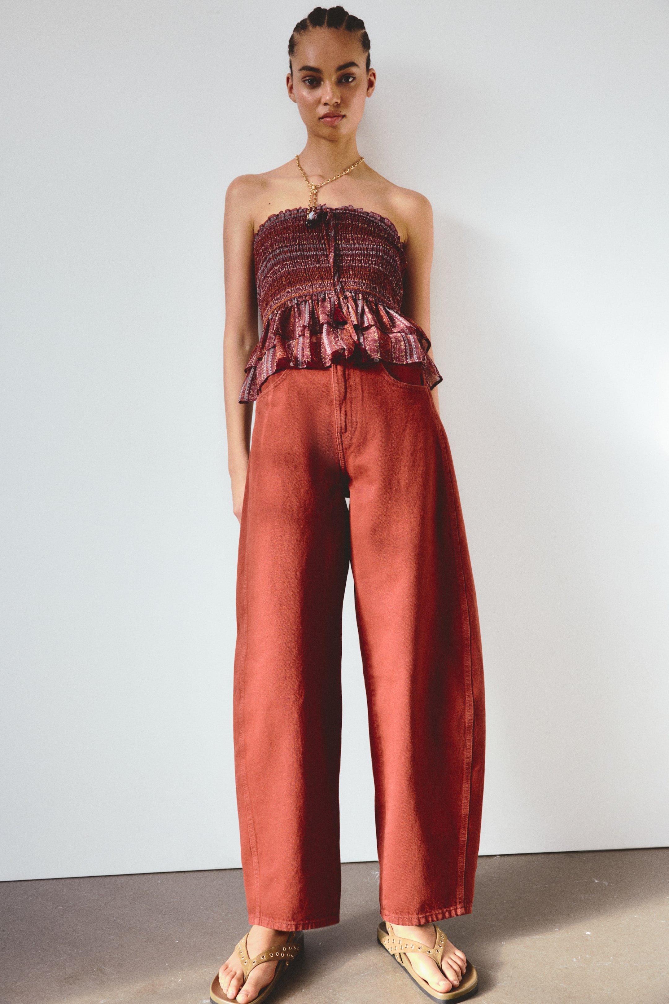 View larger image: A strapless smocked top with a ruffled peplum hem, showcasing a reddish-brown subtle pattern and a central tie detail with tassels, is displayed from a straight-on view. It is styled with high-waisted, wide-leg pants in solid burnt orange, a gold chain necklace with a round pendant, and light tan thong sandals adorned with studs.