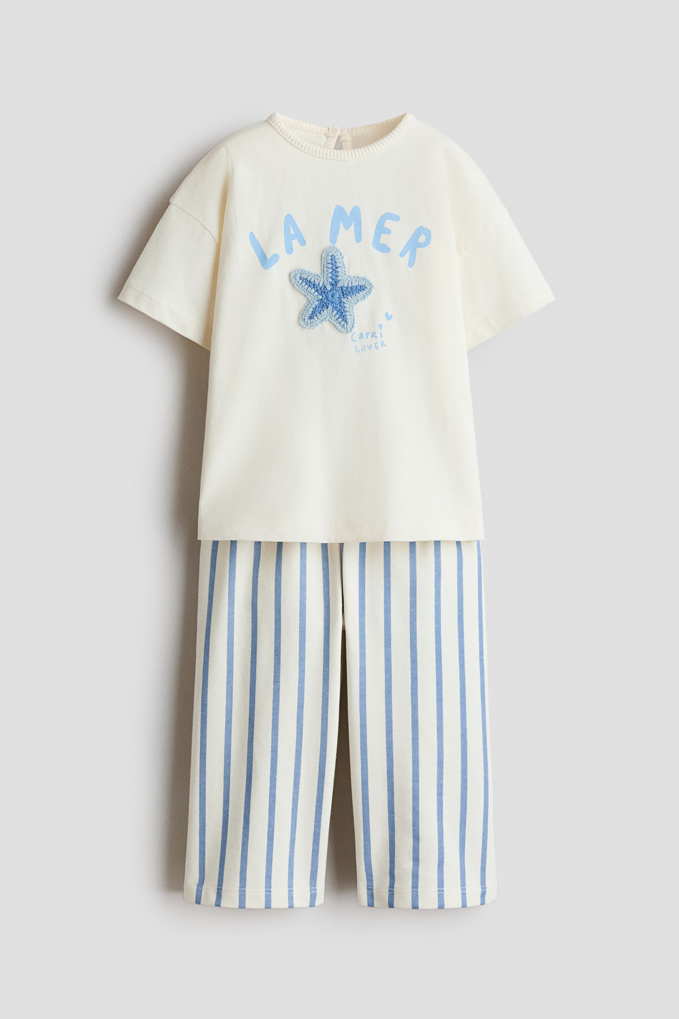 Grotere afbeelding bekijken: Een tweedelige outfit bestaande uit een gebroken wit T-shirt met korte mouwen, ronde hals en bijpassende gestreepte broek, allemaal naar voren gericht. Het T-shirt is voorzien van "LA MER" in lichtblauwe letters, een lichtblauwe gehaakte zeester-applicatie en de tekst "CAPRI LOVER", met een kleine knoopsluiting zichtbaar aan de achterkant van de halslijn. De broek toont crèmekleurige stof met lichtblauwe verticale strepen.