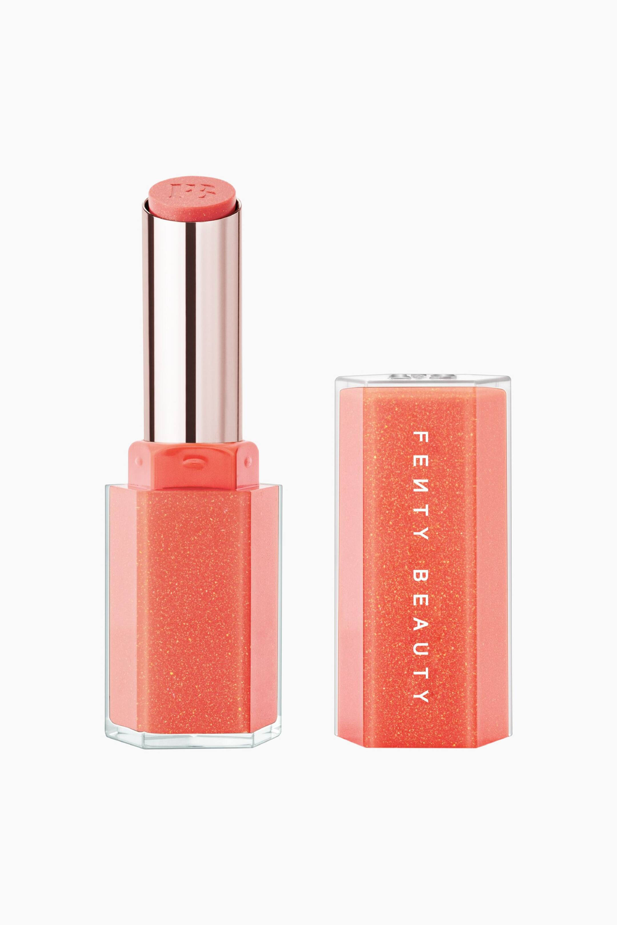 Näytä suurempi kuva: Gloss Bomb Stix High-shine Shimmer Gloss Stick seisoo pystyssä irrotetun korkkinsa vieressä, esitellen läpikuultavaa, korallinoranssia kuusikulmaista pohjaa, jossa on kultainen helmiäishohto. Auki oleva applikaattori paljastaa metallinhohtoisen ruusukultaisen sisäputken, jossa on korallinsävyinen huulikiiltotikku ja siihen kohokuvioitu "FB"-logo, kun taas pystyssä oleva korkki näyttää "FENTY BEAUTY" -logon pystysuunnassa valkoisin kirjaimin.