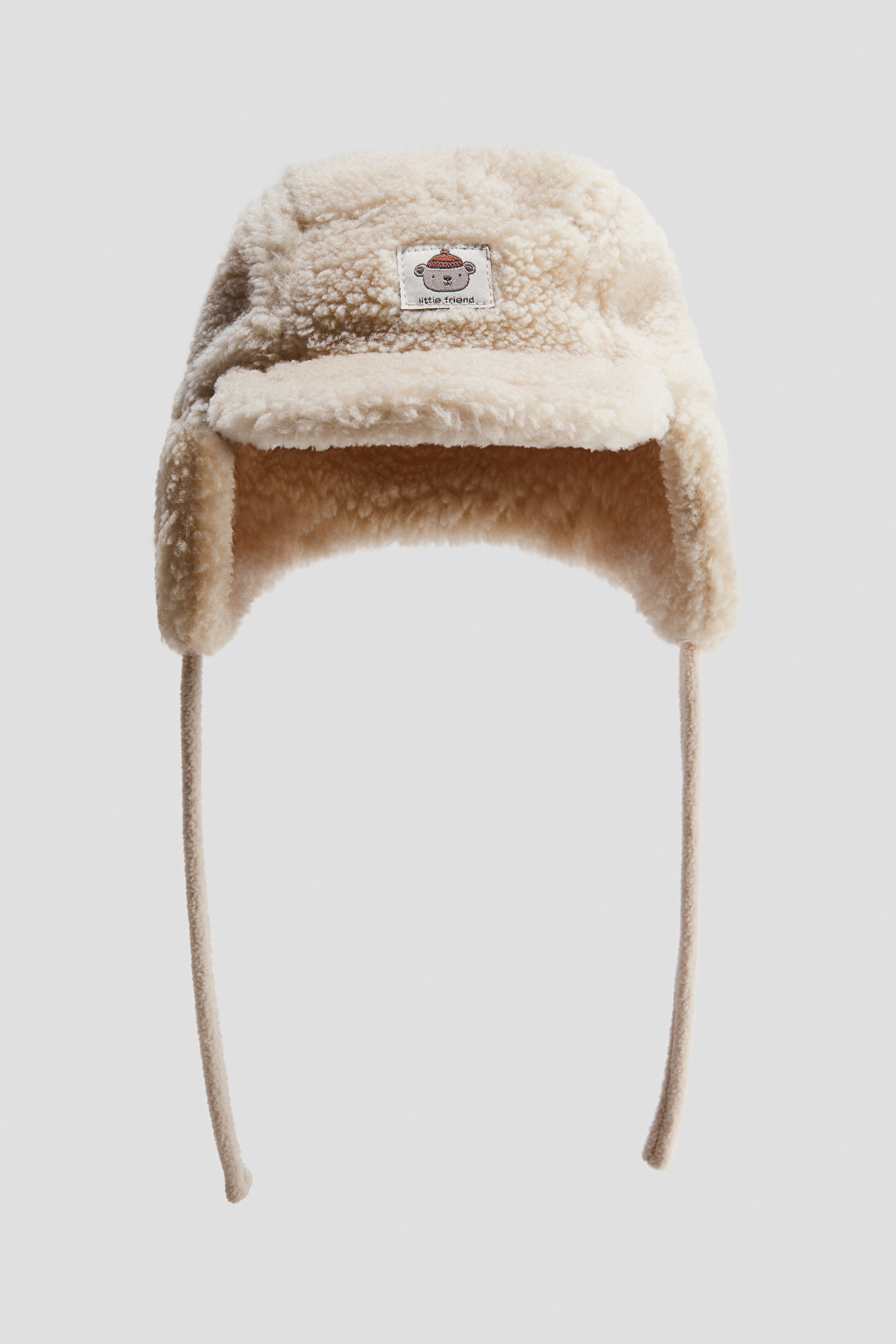 Agrandir l'image: Une casquette à cache-oreilles est présentée de face, confectionnée dans une matière teddy douce de couleur crème. Elle est dotée de longues attaches pendantes depuis les cache-oreilles et d’une petite étiquette rectangulaire avec un motif de singe et le texte « little friend » cousue sur l’avant.