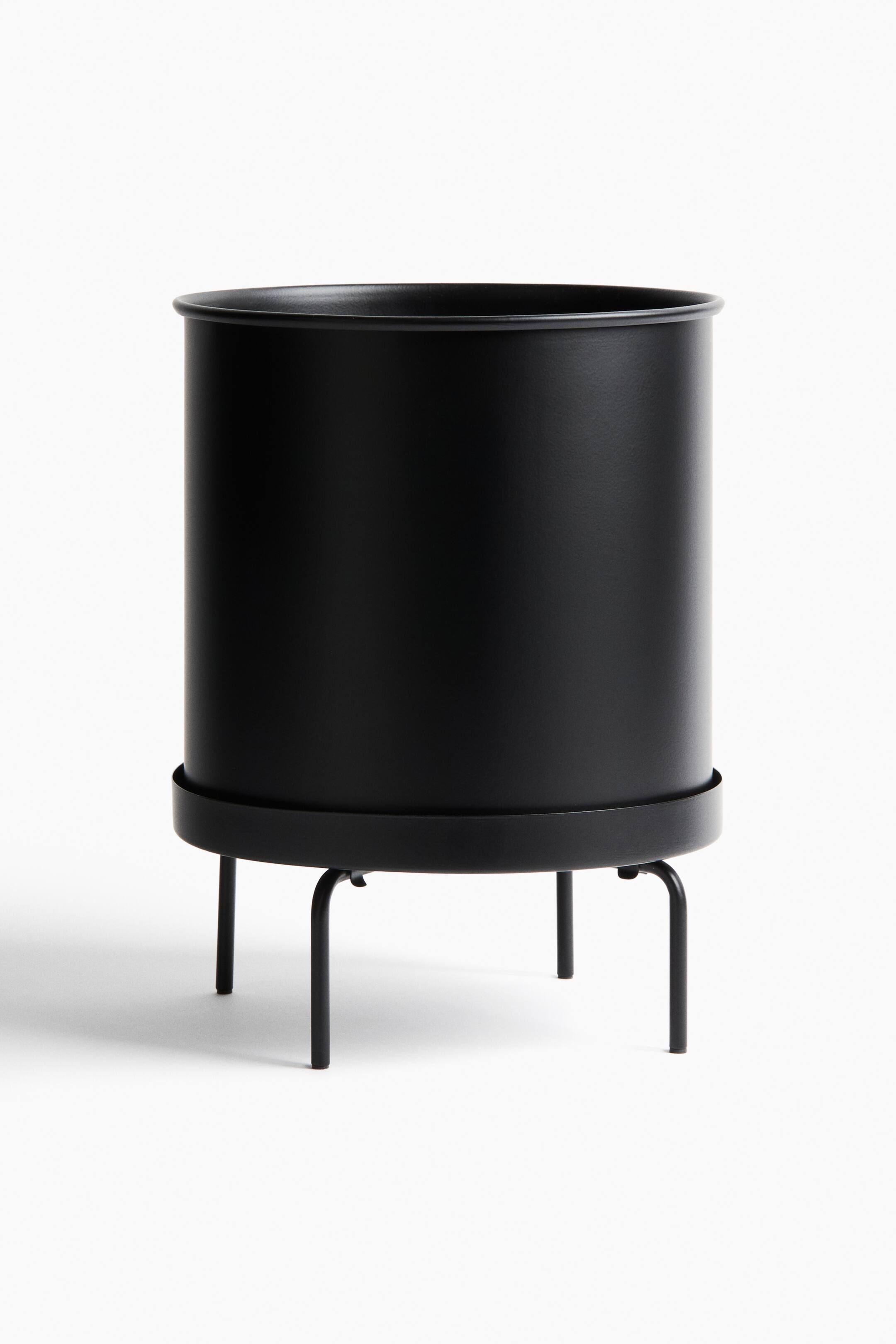 View larger image: A black cylindrical planter is presented from the front, resting on a matching black metal stand. The planter is showing a rolled rim at its top, and the stand is supporting the planter with a circular base and four short, curved legs.