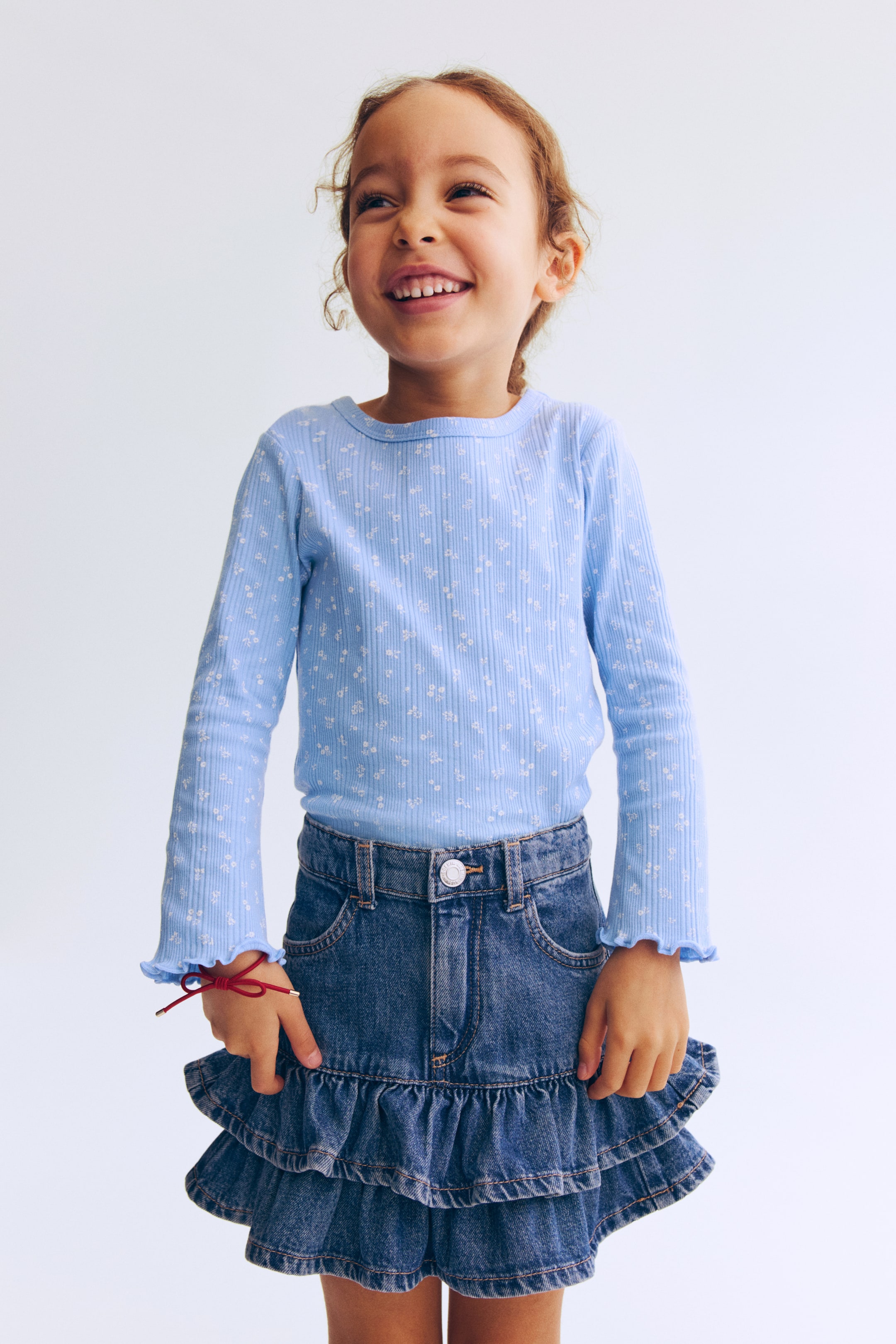 Zobacz większy obraz: Przód jasnoniebieskiego, prążkowanego topu z dżerseju zdobi drobny, biały motyw kwiatowy na całej powierzchni. Model ma okrągły dekolt wykończony prążkowaną lamówką oraz długie rękawy zakończone falbankami obszytymi ściegiem overlock przy mankietach.