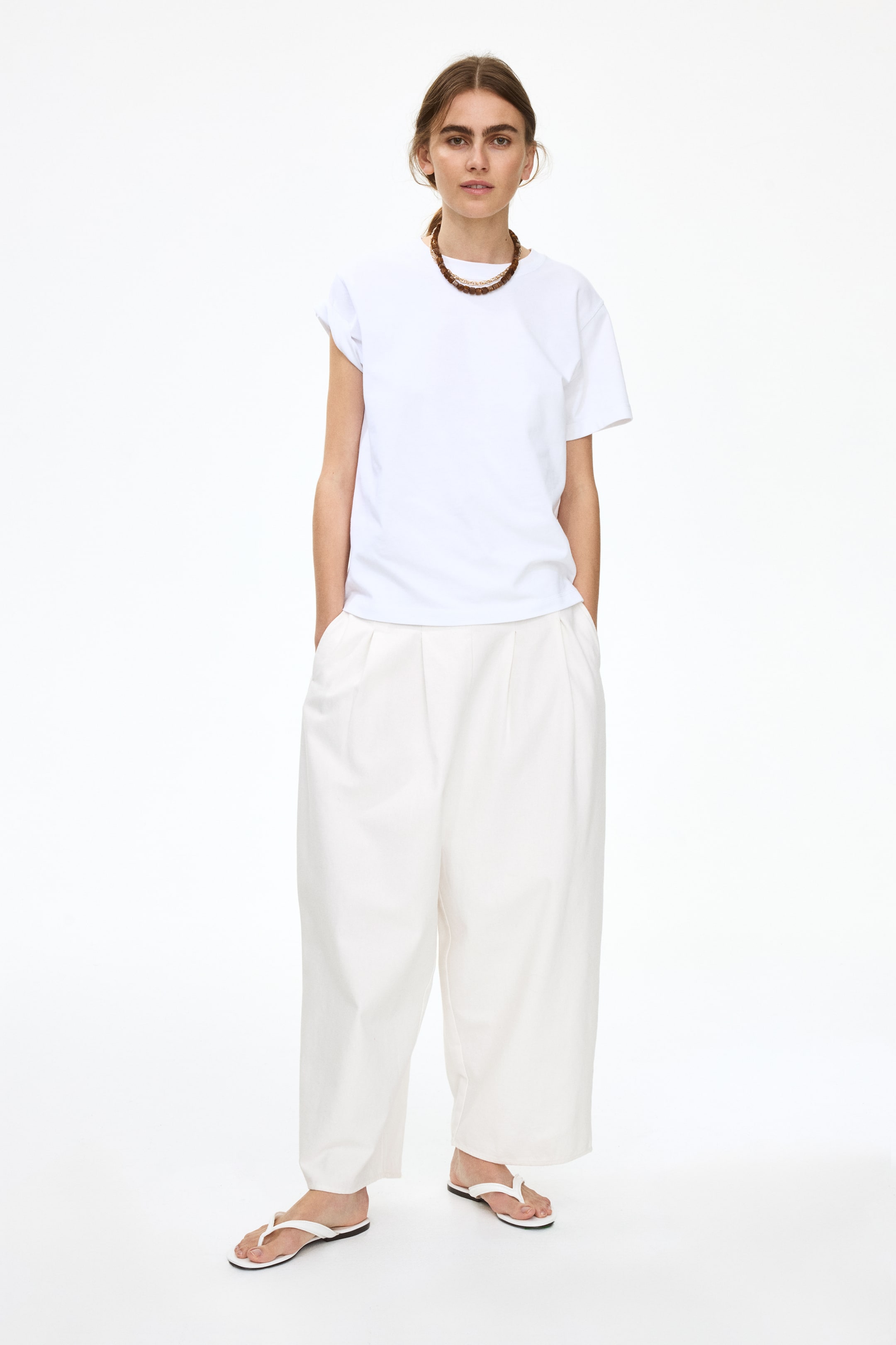 Agrandir l'image: Un t-shirt blanc à col rond avec des manches retroussées est porté, face à l'avant, associé à un pantalon large à plis de couleur claire et des tongs blanches. Un collier multi-rangs, avec une chaîne dorée et des perles brunes, orne le cou.
