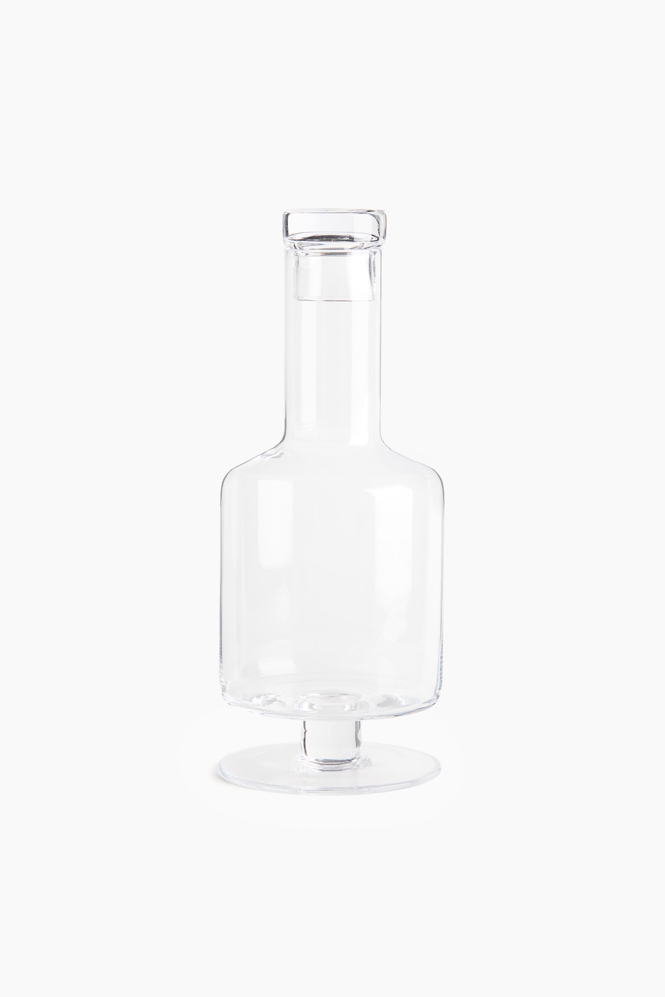 Ver imagen más grande: Un decantador de vino con base, visto de frente, presenta una construcción de vidrio soplado a mano, transparente, con bordes suavemente redondeados. Cuenta con un cuello estrecho, con un tapón de vidrio a juego descansando en su interior, y una base ancha y plana.