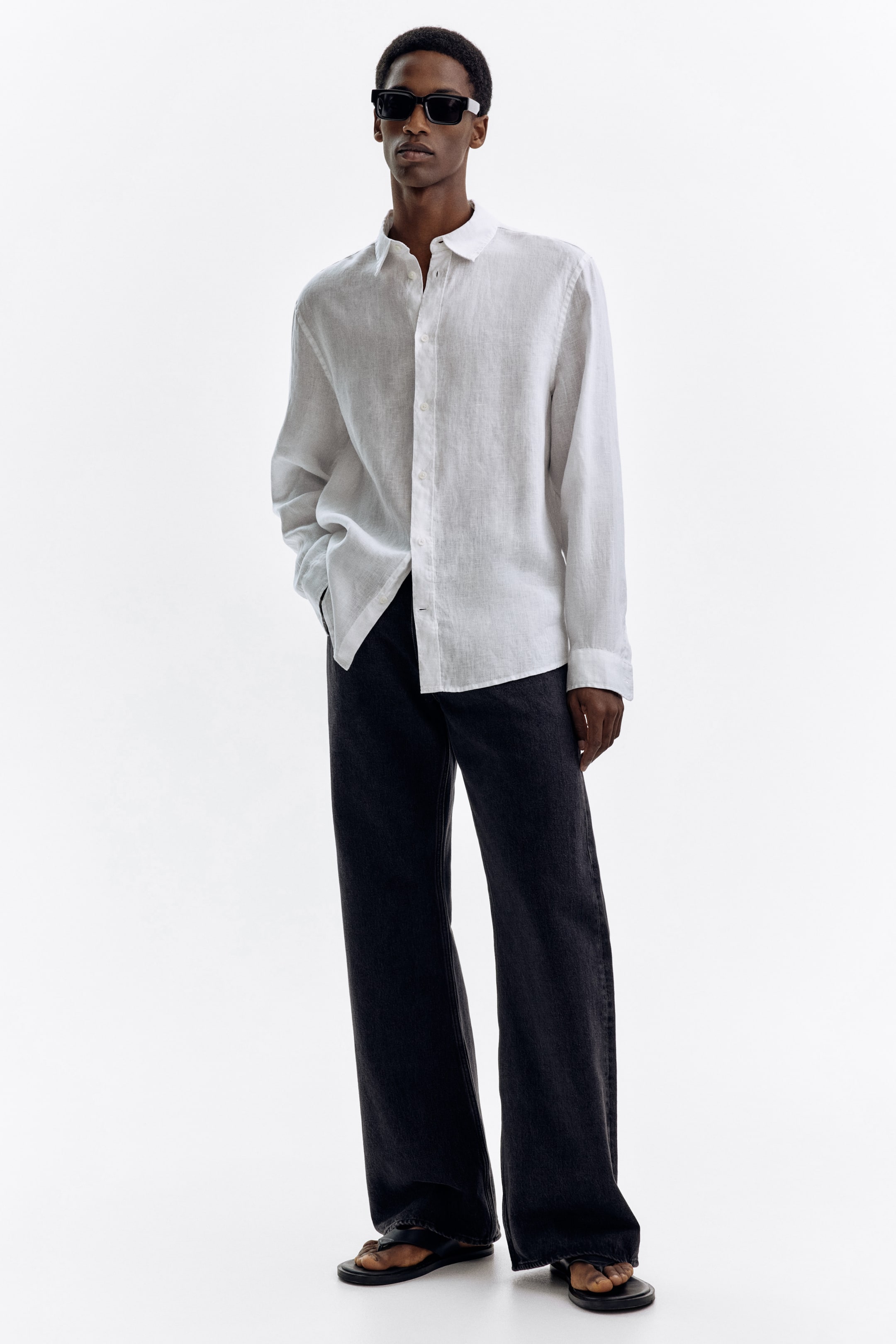 Zobacz większy obraz: Biała koszula z długim rękawem, z charakterystycznym splotem i klasycznym kołnierzykiem, noszona jest na wierzchu ciemnych, szerokich spodni. Spodnie mają luźny krój i są stylizowane z czarnymi sandałami typu japonki oraz czarnymi prostokątnymi okularami przeciwsłonecznymi.