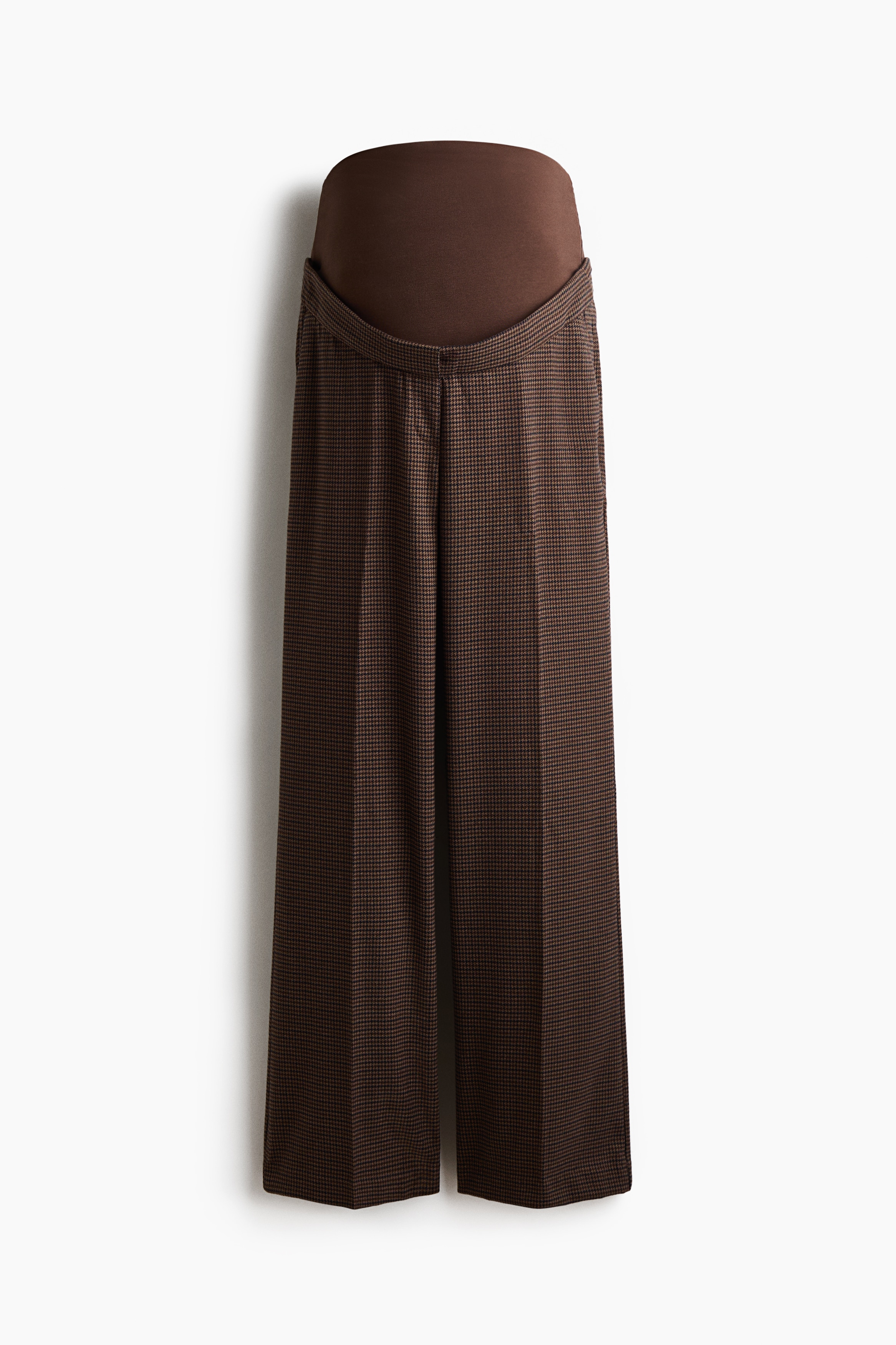Bruine broek van tweedstof met patroon, met bootcut-pijpen en strakke plooien langs de voorkant, gedragen door het model. De broek is gestyled met een bijpassende blazer, vastgehouden met een dunne zwarte riem boven de taille, en zwarte loafers.