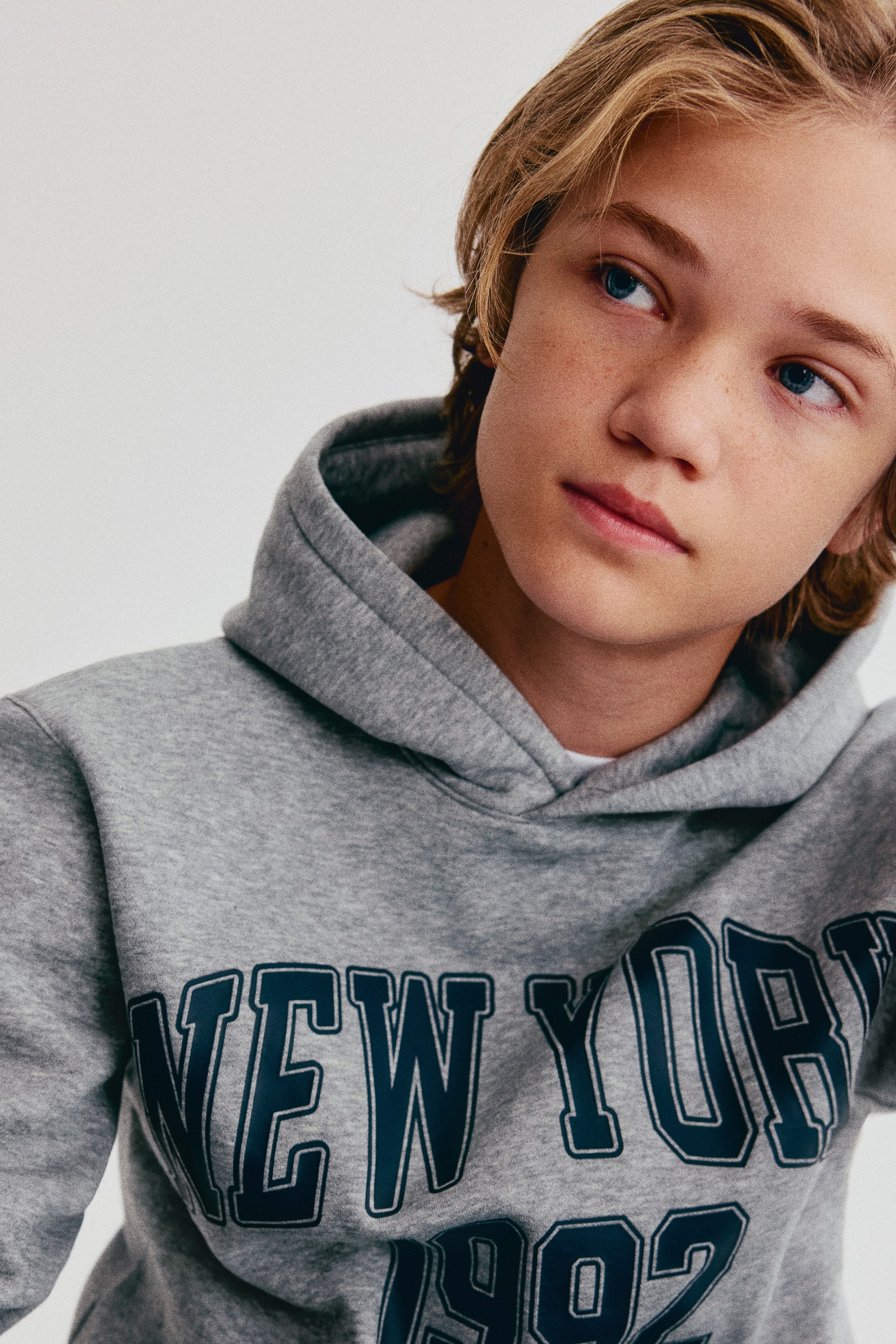 Größeres Bild ansehen: Ein grauer Hoodie aus Sweatstoff ist von vorne zu sehen und verfügt über eine gefütterte Kapuze mit Wickel-Ausschnitt sowie einen dunkelblauen College-Print „NEW YORK 1992“ auf der Brust.