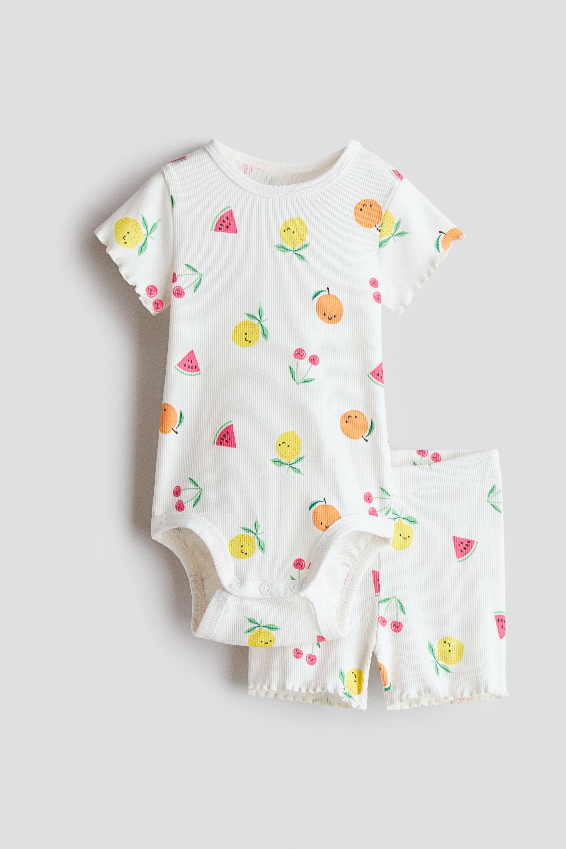View larger image: A white ribbed cotton jersey set, featuring a bodysuit and cycling shorts, showcases its front adorned with vibrant fruit patterns, including lemons, oranges, watermelon slices, cherries, and small pink flowers. The bodysuit boasts a trimmed neckline, short ruffled sleeves, and snap closures at the crotch, while the cycling shorts display ruffled hems.