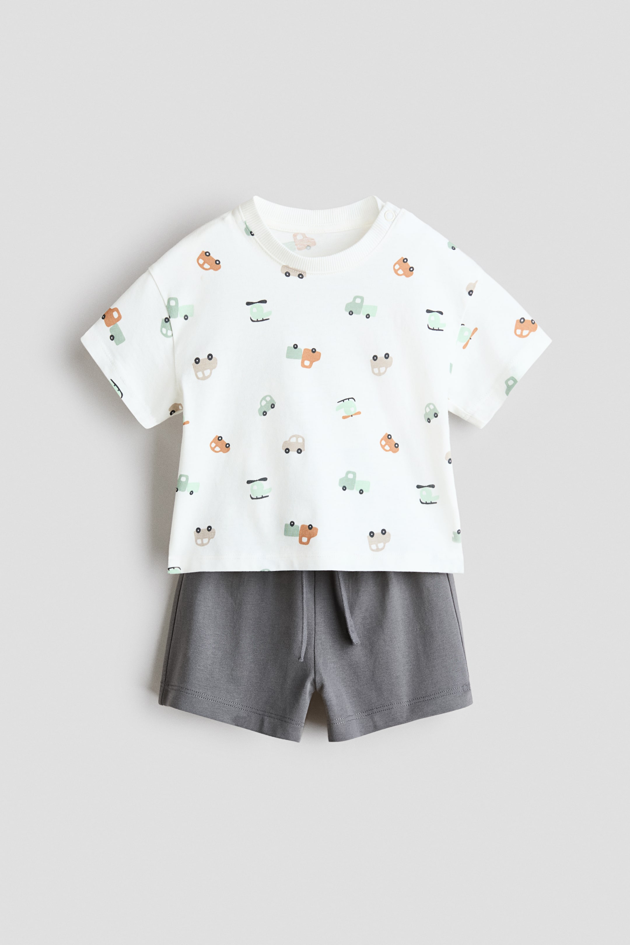 Xem ảnh lớn hơn: Bộ đồ cotton 2 mảnh được trưng bày từ phía trước, bao gồm áo thun trắng ngắn tay với họa tiết phương tiện đầy màu sắc, bao gồm ô tô, xe tải và máy bay trực thăng, cùng cổ tròn bo gân có nút bấm ở vai. Quần short màu xám đậm hoàn thiện bộ đồ, có cạp chun với dây rút có thể nhìn thấy.