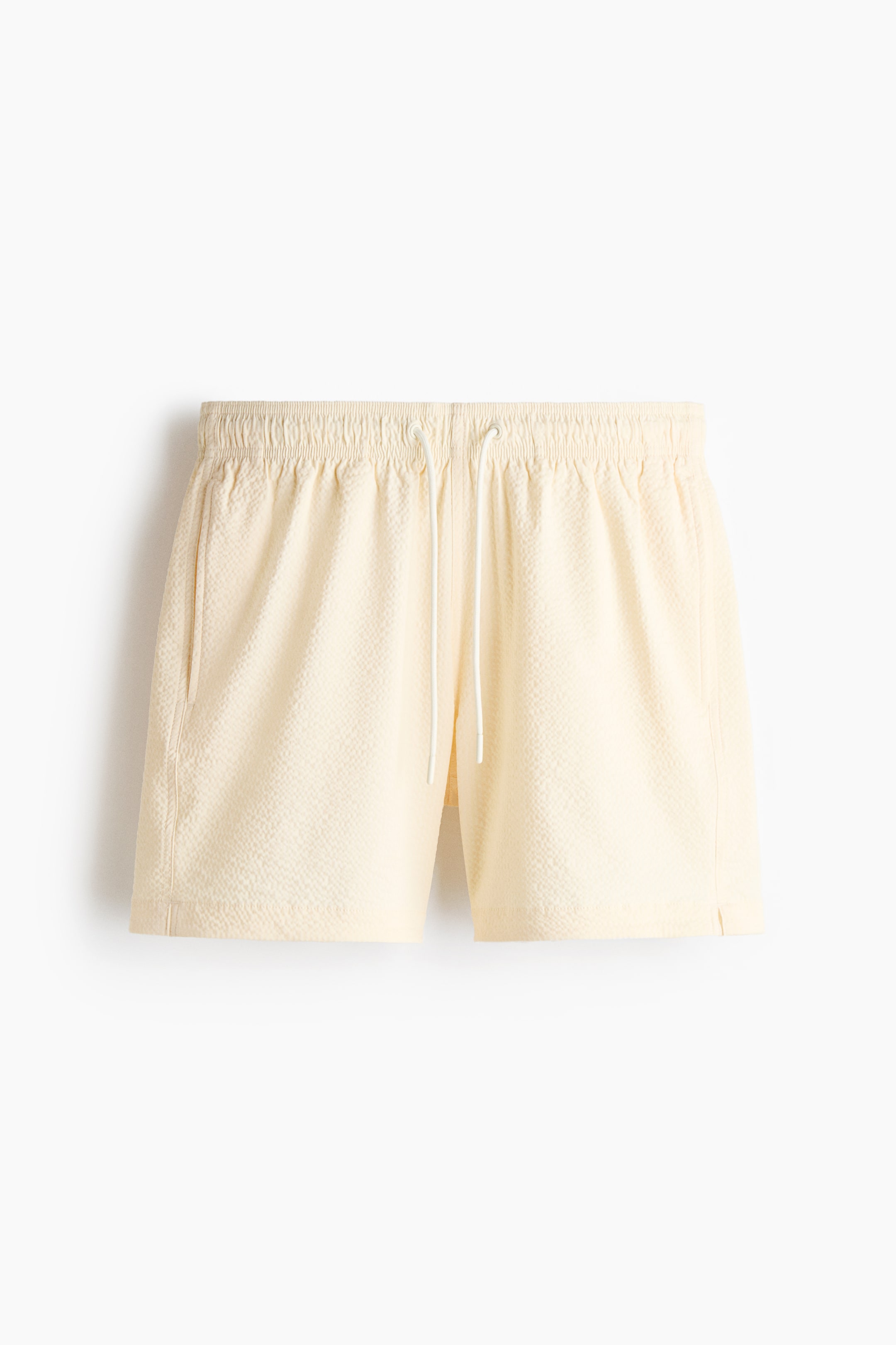 View larger image: Mid-length pale yellow seersucker swim trunks are shown from the FRONT, featuring an elastic waistband with a white drawstring, side pockets, and small slits at the side hems.
