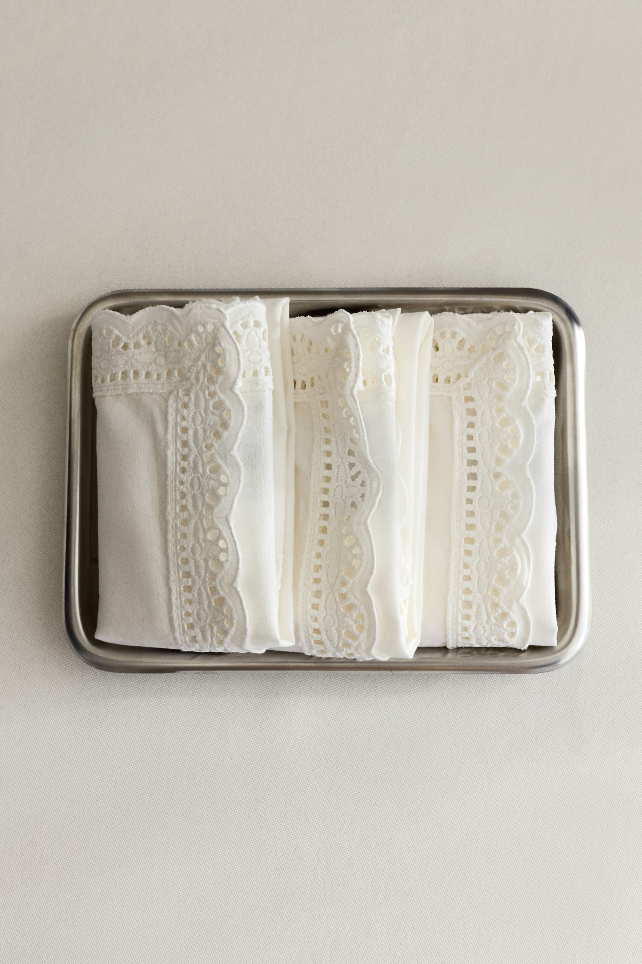 View larger image: White folded fabric items, each featuring a wide scalloped eyelet lace trim, are resting side by side in a rectangular metal tray.