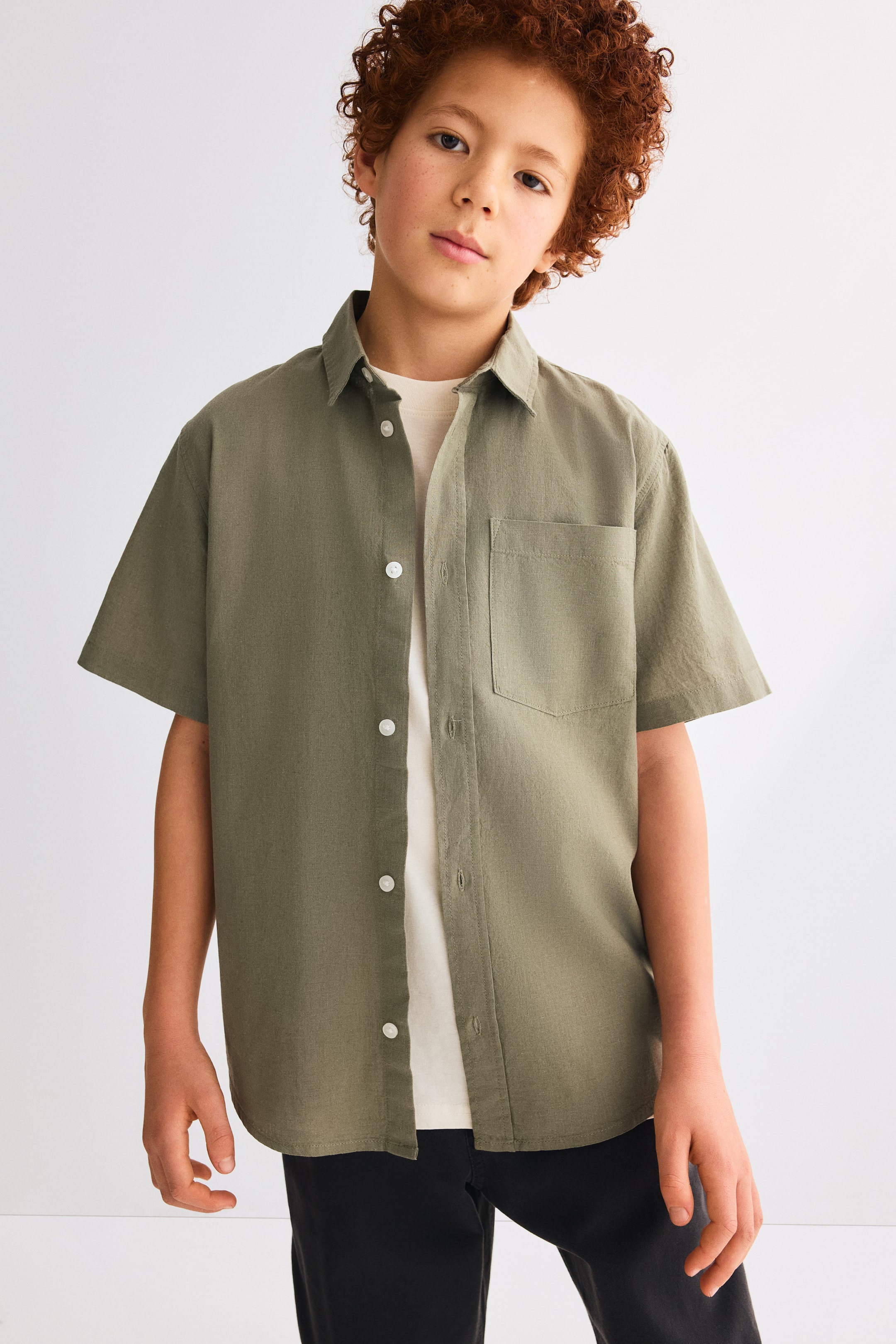 Ver imagem maior: Uma camisa verde-oliva de manga curta, voltada para a frente, apresenta uma gola padrão, botões brancos numa frente abotoada e um bolso único no peito do lado esquerdo, sendo usada aberta sobre uma t-shirt de cor clara.