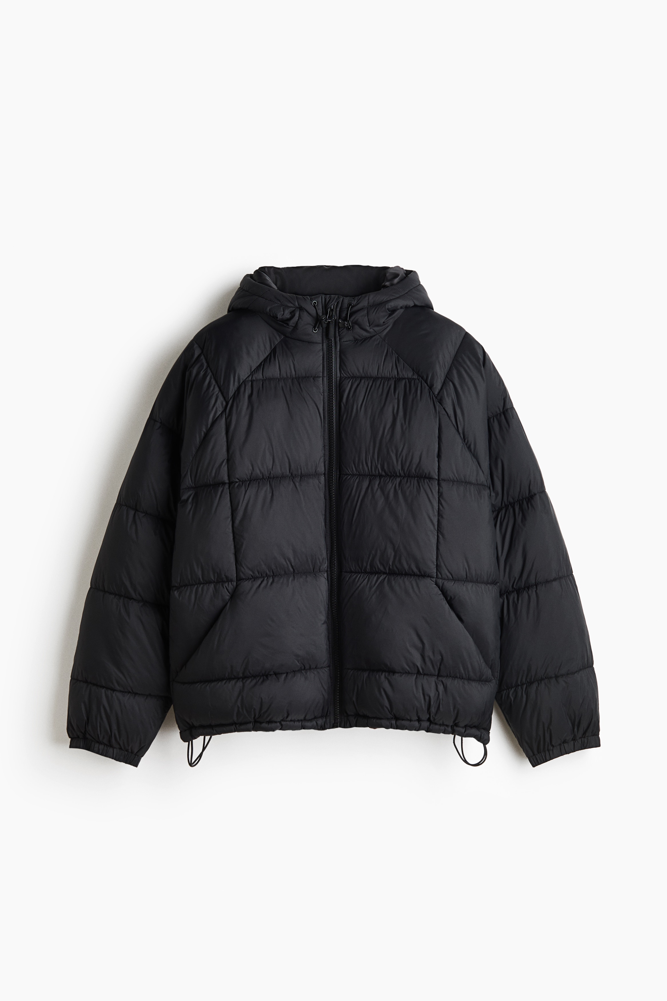 A black padded puffer jacket featuring a quilted weave, a stand collar, and a hood is being worn by the model. The model is standing with hands in the front pockets, styling the jacket with wide-leg grey jeans and a black crossbody strap.