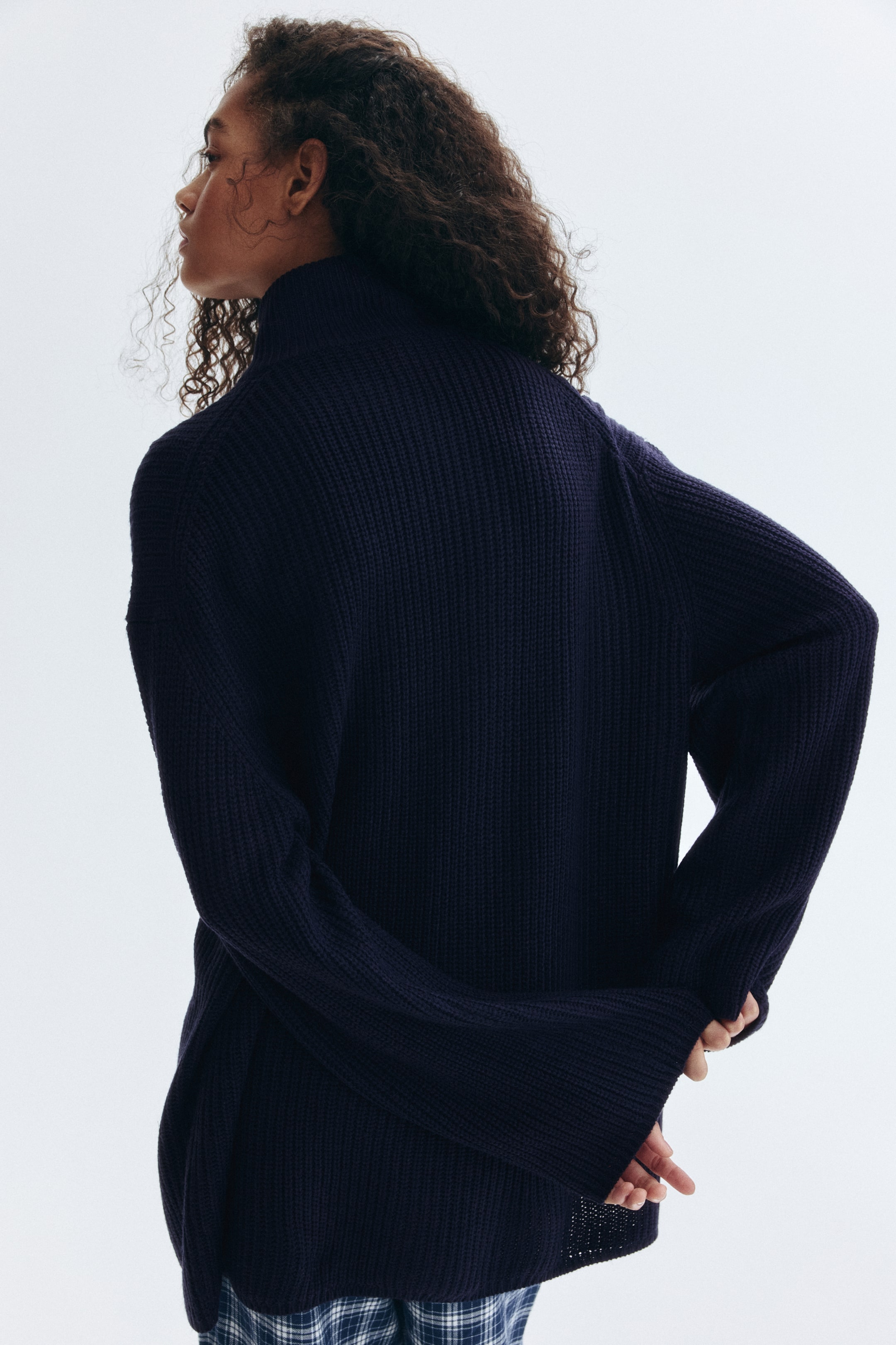 View larger image: A dark blue ribbed knit turtleneck sweater is being worn by the model. Its long, oversized sleeves are covering the model's hands, which are clasped behind their back, while plaid pants are visible underneath.
