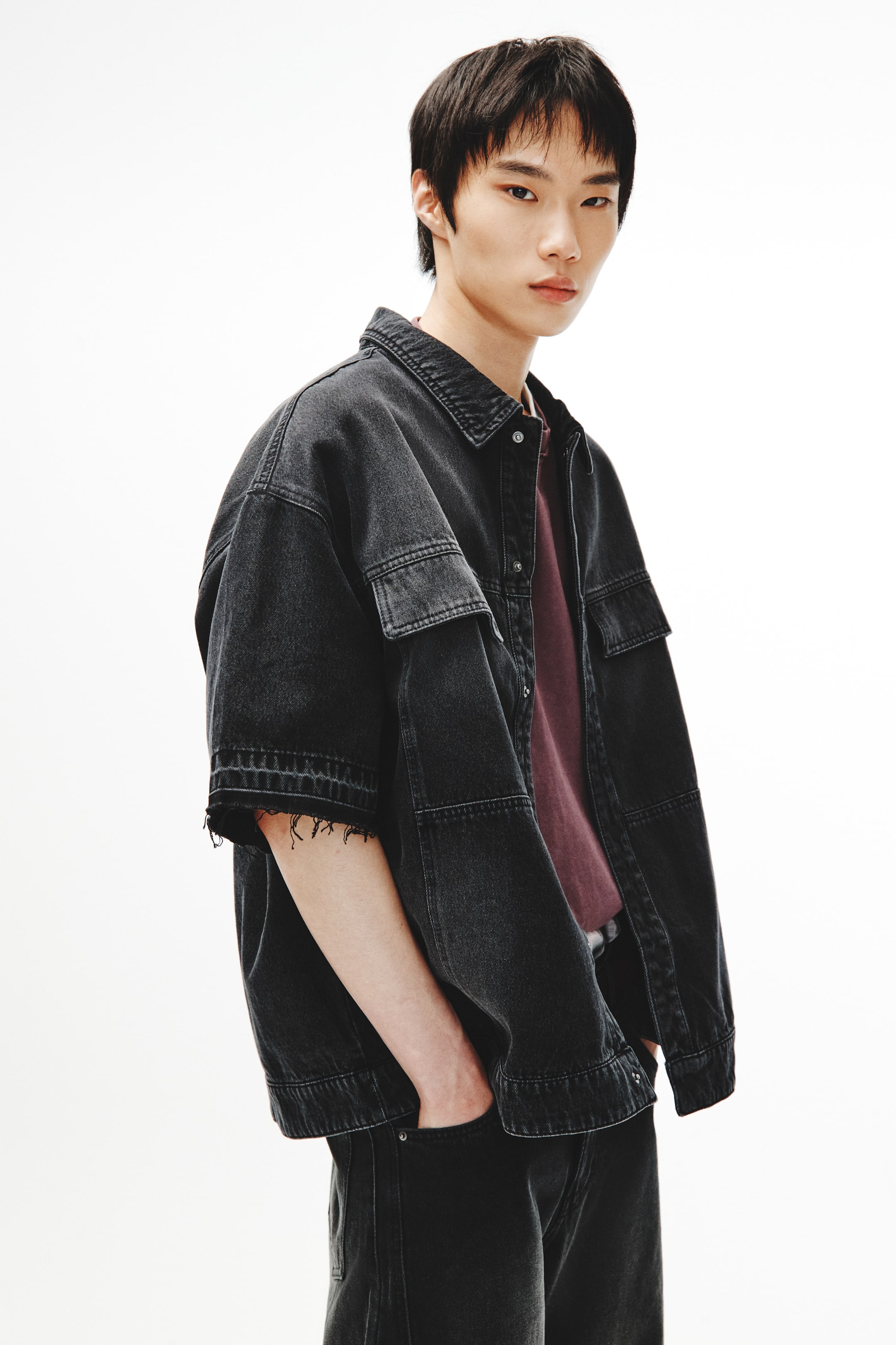 Xem ảnh lớn hơn: Một chiếc áo sơ mi ngắn tay denim tối màu đang được giới thiệu, hơi nghiêng về bên phải. Nó có cổ nhọn, hàng cúc phía trước, hai túi nắp lớn ở phần ngực trên và hai túi vá bổ sung ở phần trước dưới. Tay áo ngắn rộng rãi với các cạnh xù xì, tua rua, và nó đang được mặc mở bên ngoài một chiếc áo thun màu nâu đỏ.
