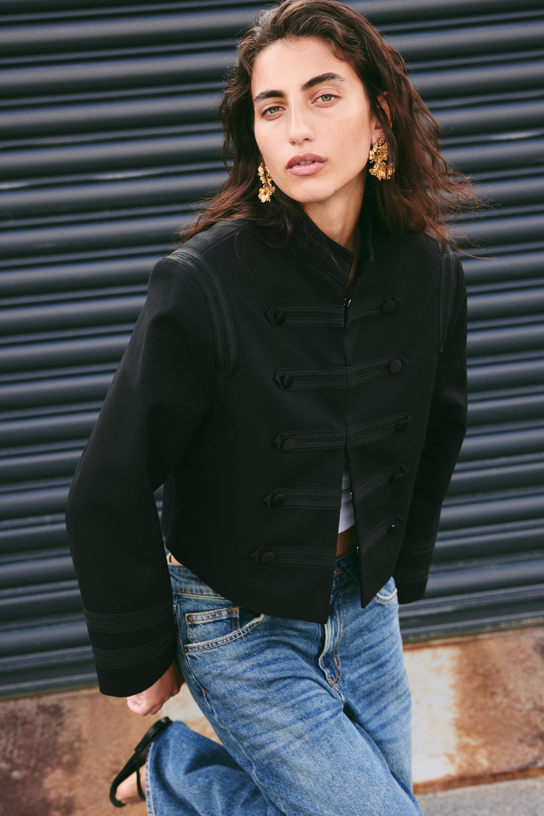 View larger image: The model is wearing a short black twill jacket featuring a stand collar, shoulder pads, and concealed hook-and-eye fastenings. Decorative trims and covered buttons are visible at the front, with additional trim detailing at the collar and cuffs. The jacket is lined, and the model has paired it with blue jeans and a white top.