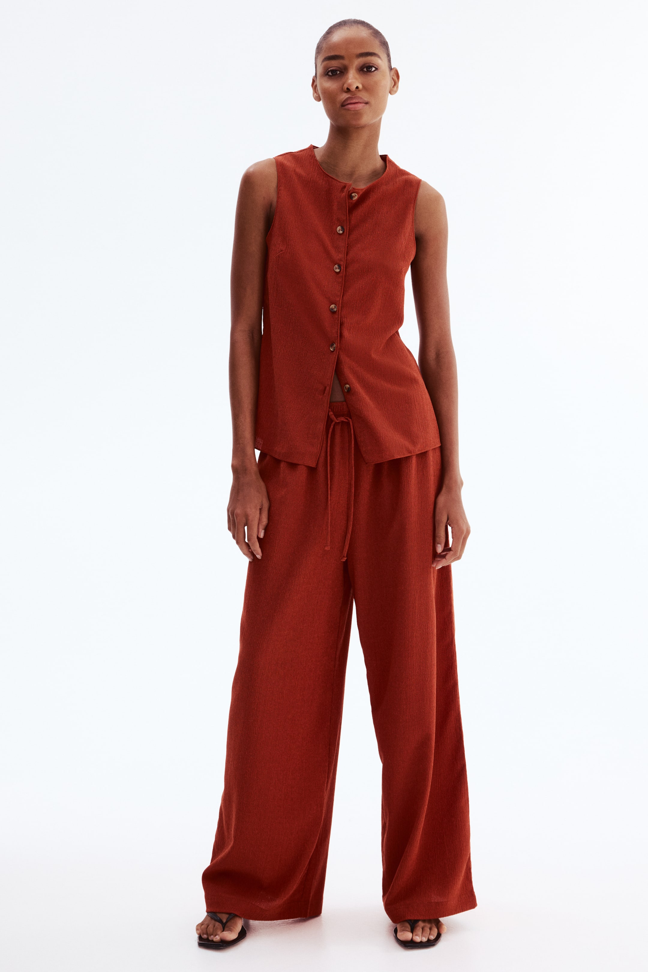 View larger image: A rust-coloured two-piece set presents a sleeveless top with a round neckline and a full front button placket, partially unbuttoned at the hem. The coordinating wide-leg pants feature a visible drawstring waist, with both pieces displaying a subtle textured weave.